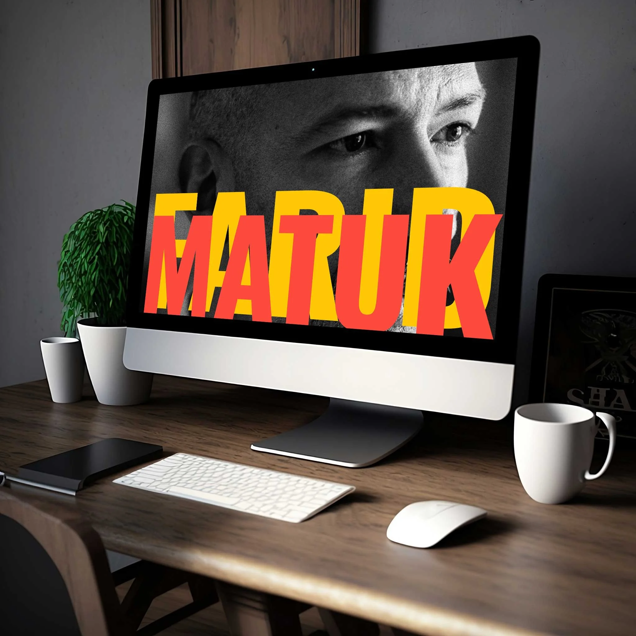 Desktop computer with large screen displaying grayscale face and bold red text "MATUK," surrounded by keyboard, mouse, coffee cup, plant, and tablet on wooden desk.