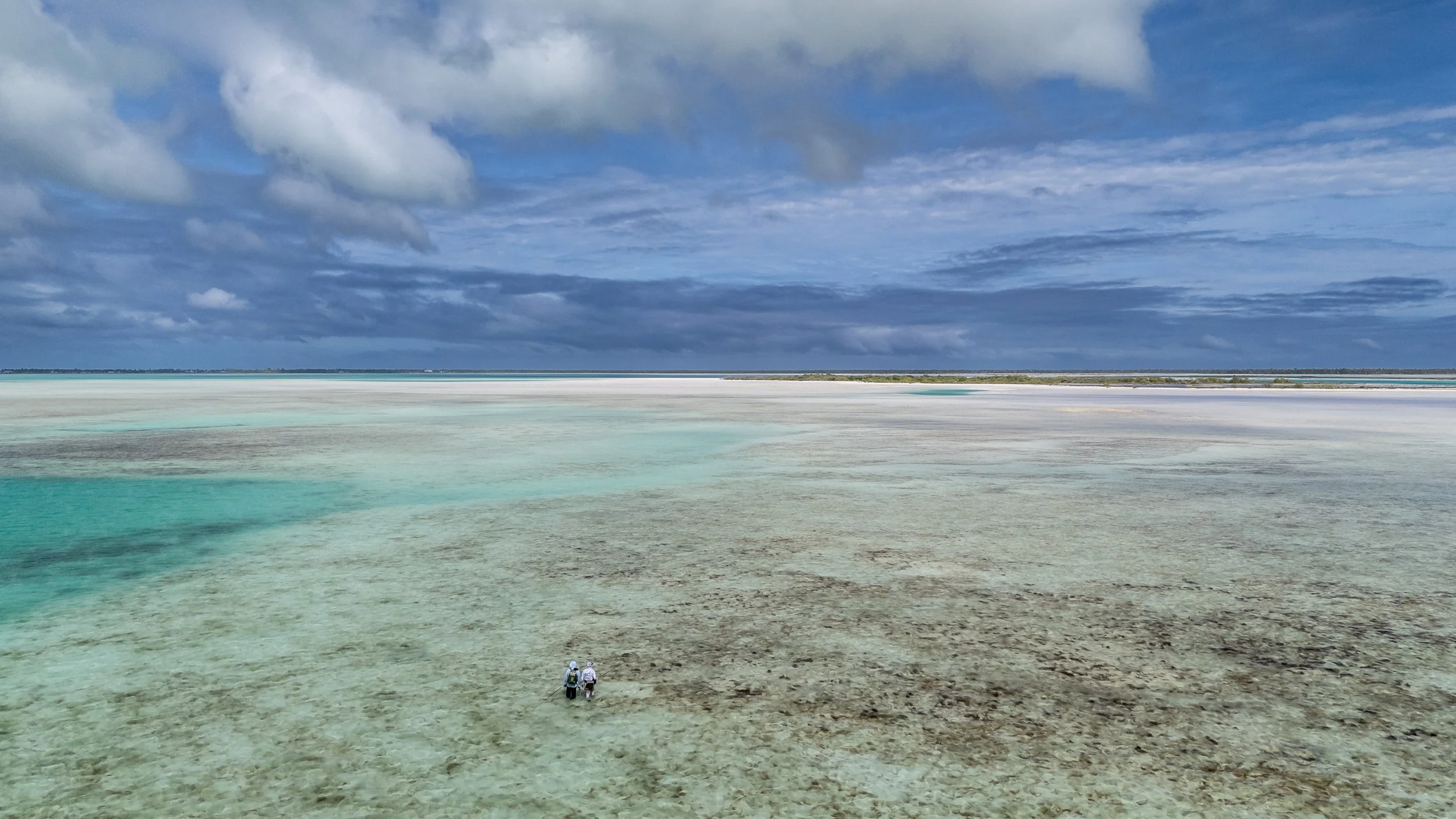 Two people walking in a shallow turquoise lagoon under a cloudy sky.