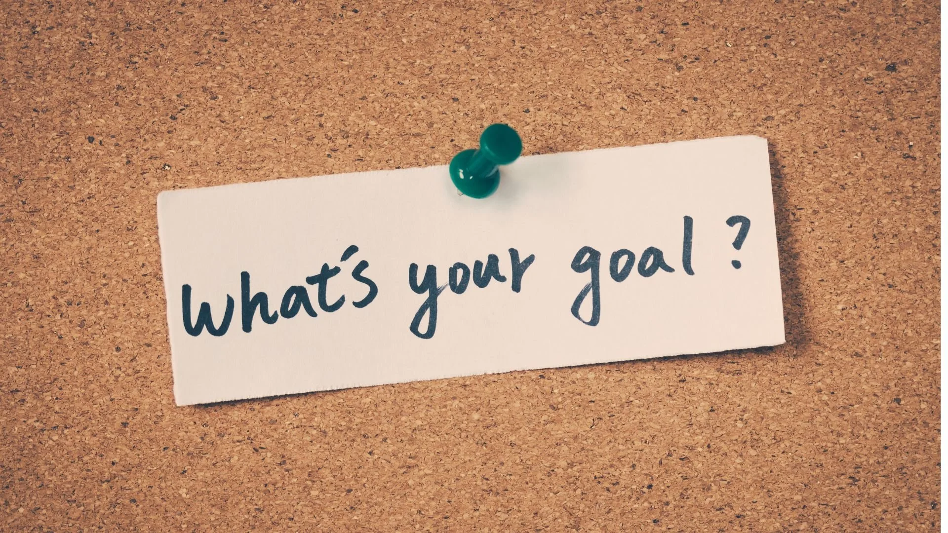 A white note pinned to a corkboard with a green push pin. The note has the handwritten question, 'What's your goal?'
