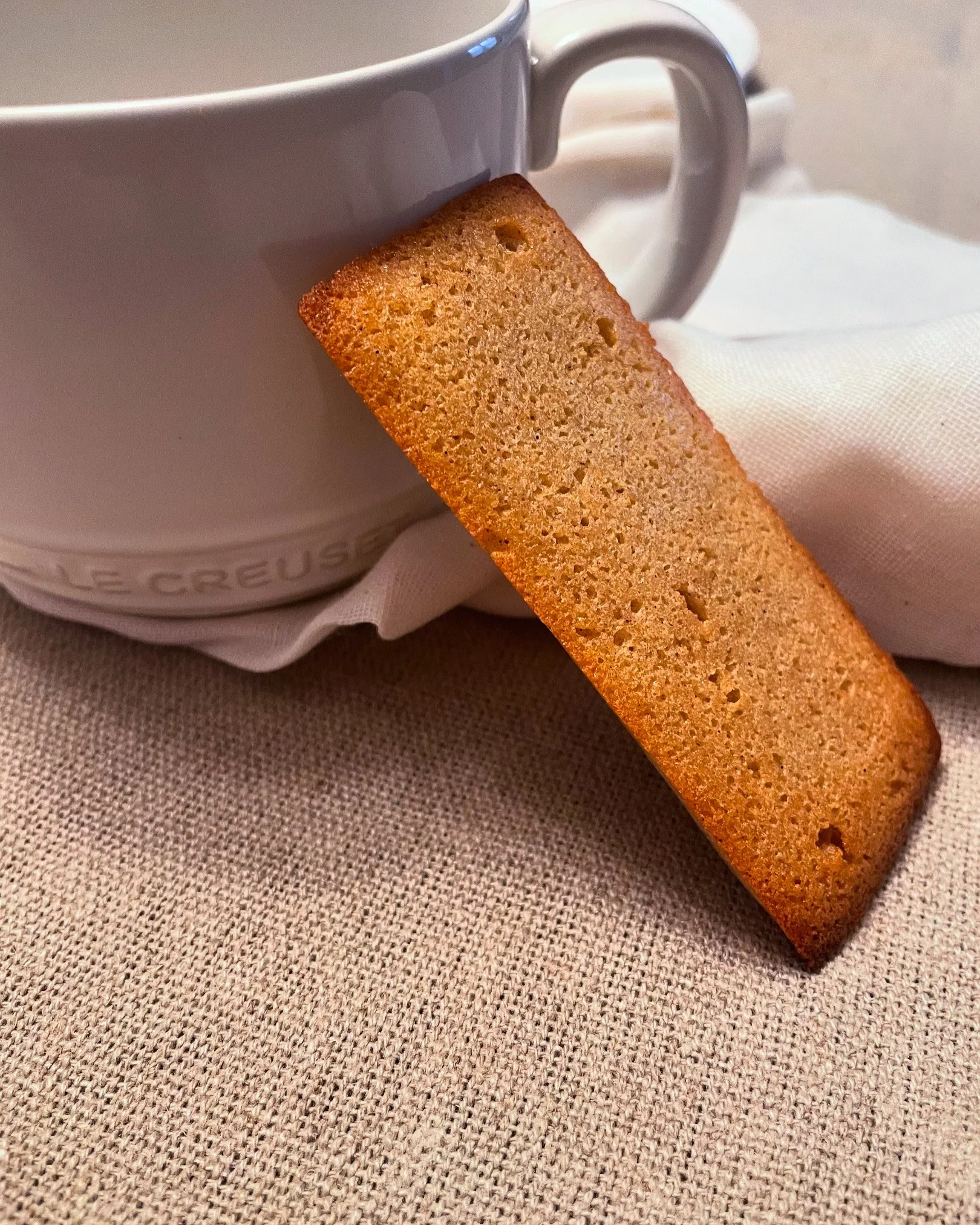 A rectangular cookie leaning against a white coffee mug on a beige textured surface.