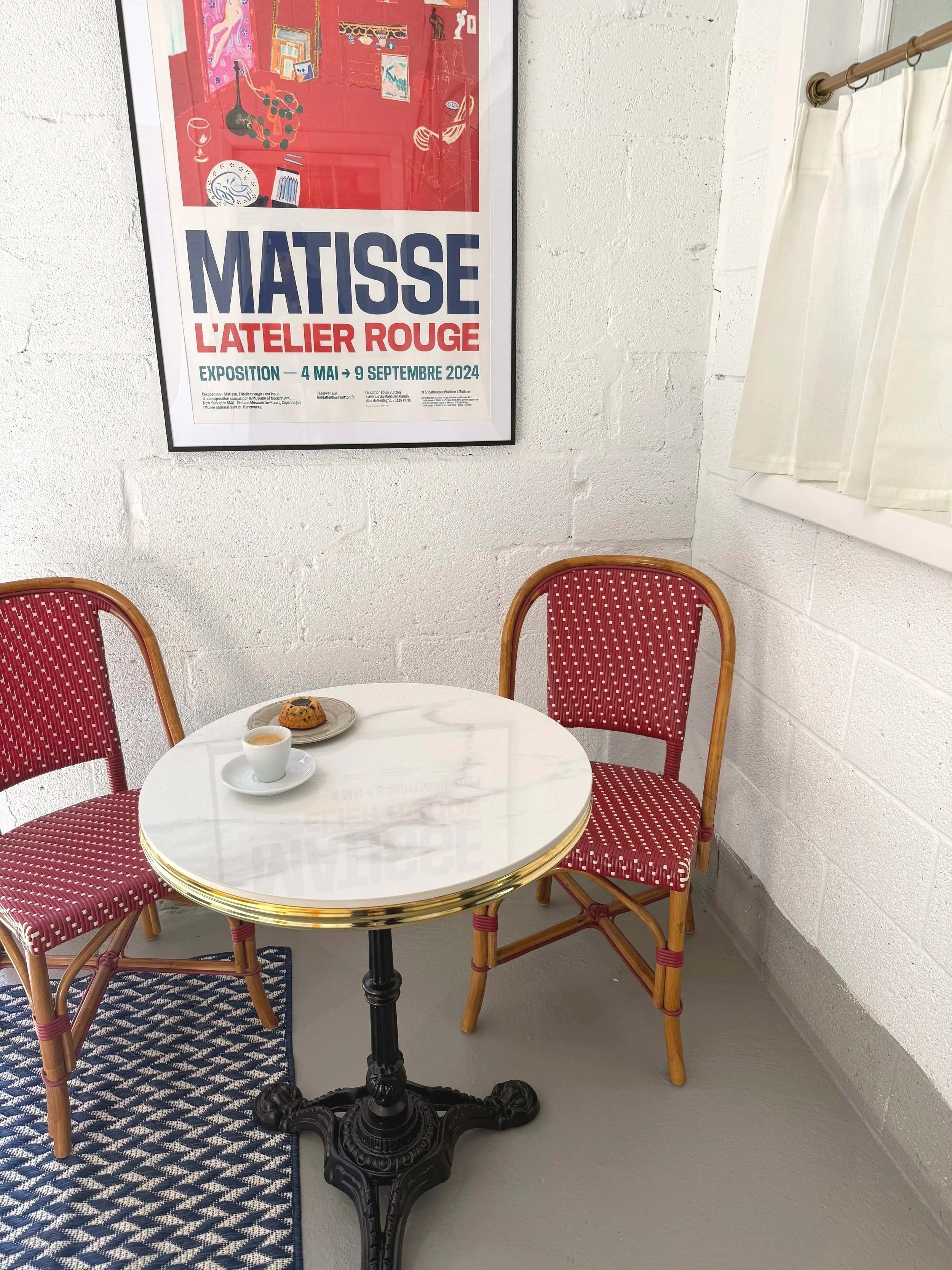 A small round table with a white marble top and ornate black base, set with a cup of coffee and a scone, surrounded by two red upholstered chairs with wooden frames, placed near a white brick wall and a window with cream curtains. A poster for a Matisse art exhibition is on the wall behind the table.