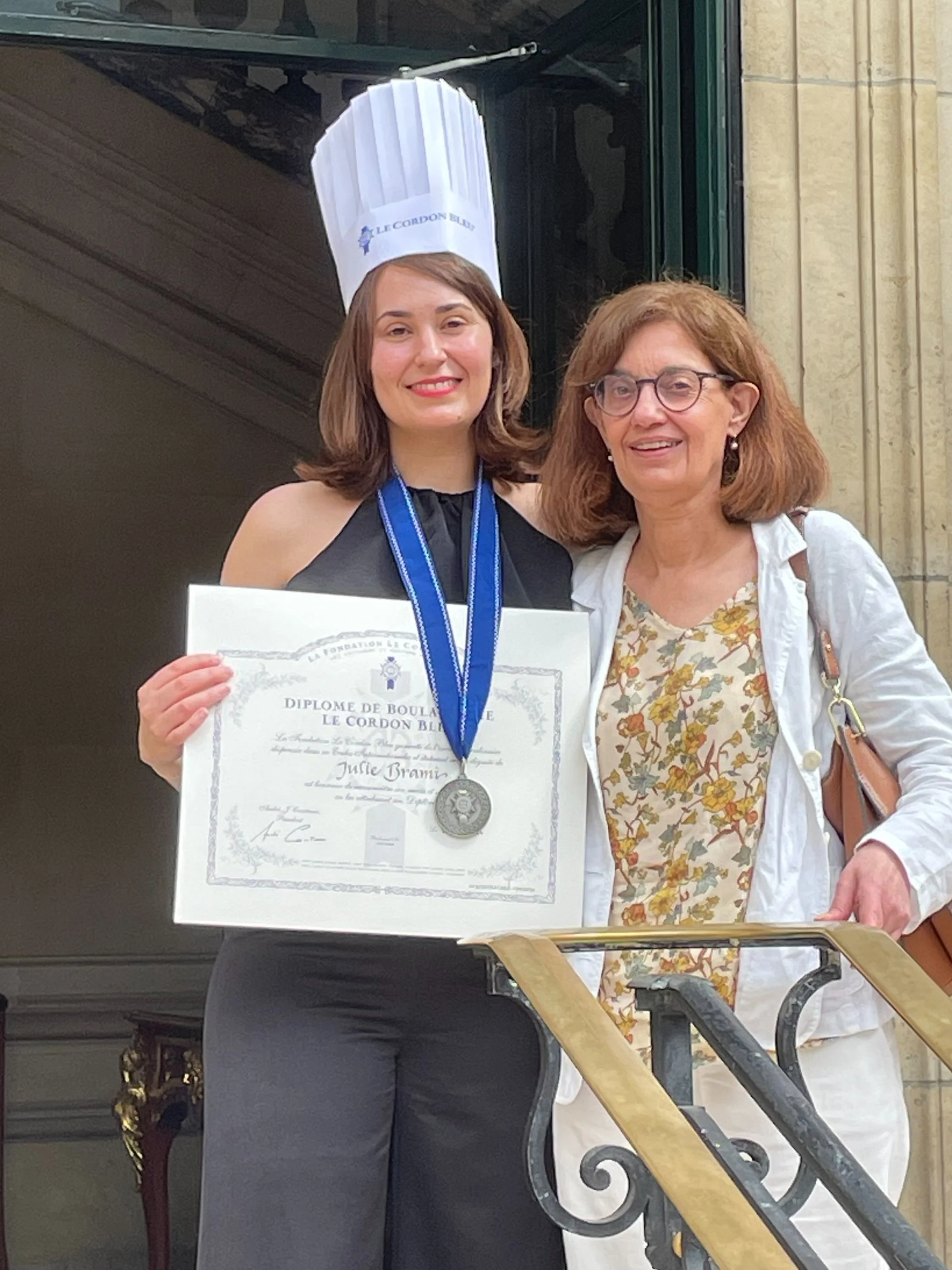 A young woman in a chef's hat and black outfit holding a diploma and medal, standing next to an older woman in white clothing on a staircase outside a building.