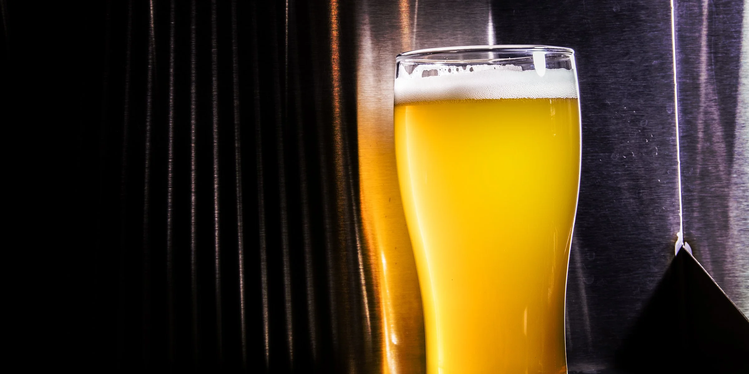 A pint glass filled with hazy, golden beer placed against a dark metal background with soft lighting and reflections.