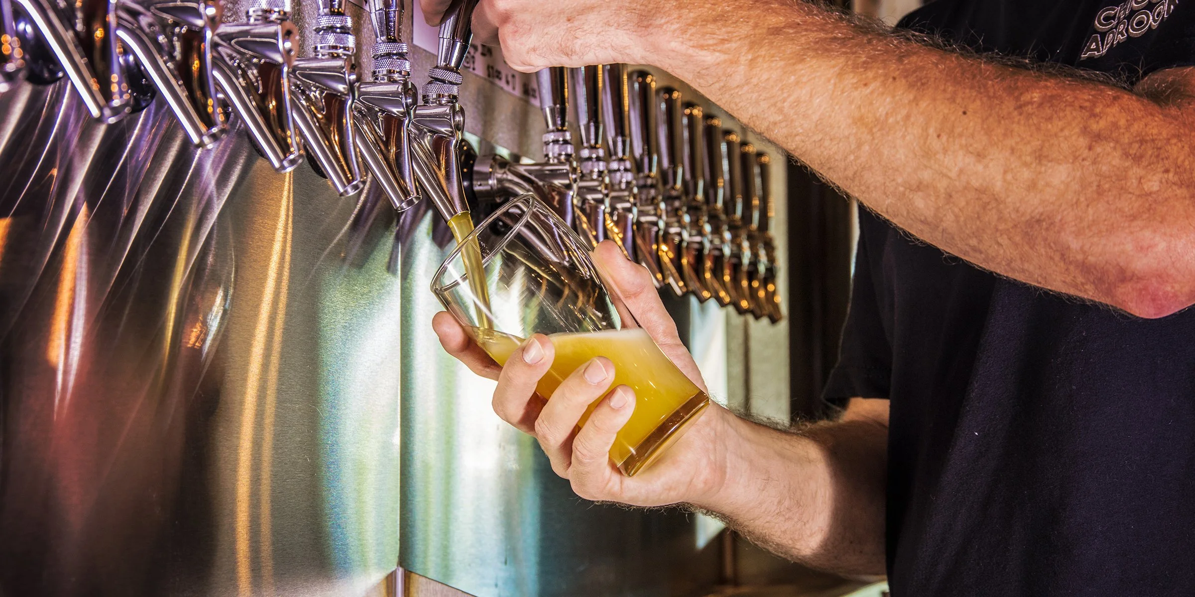 Person pouring beer from a tap into a glass at a bar, with multiple chrome beer taps visible.