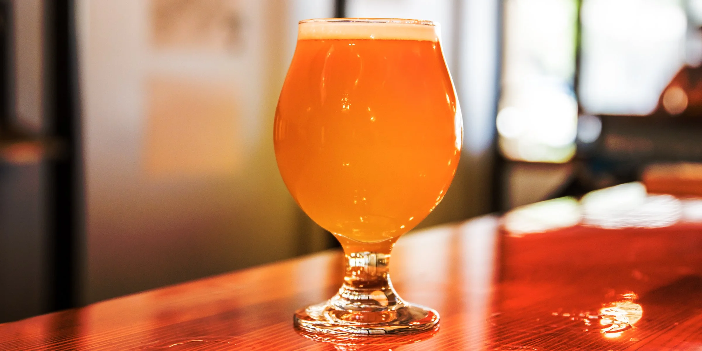 A half-full goblet of amber-colored beer on a wooden bar counter with a blurred background.