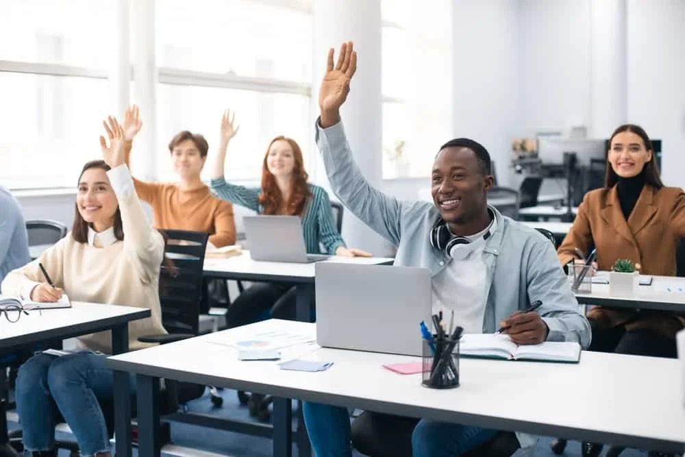 A diverse group of six young adults sitting in a classroom or meeting room, raising their hands to participate, with some taking notes and working on laptops, in a bright well-lit space.