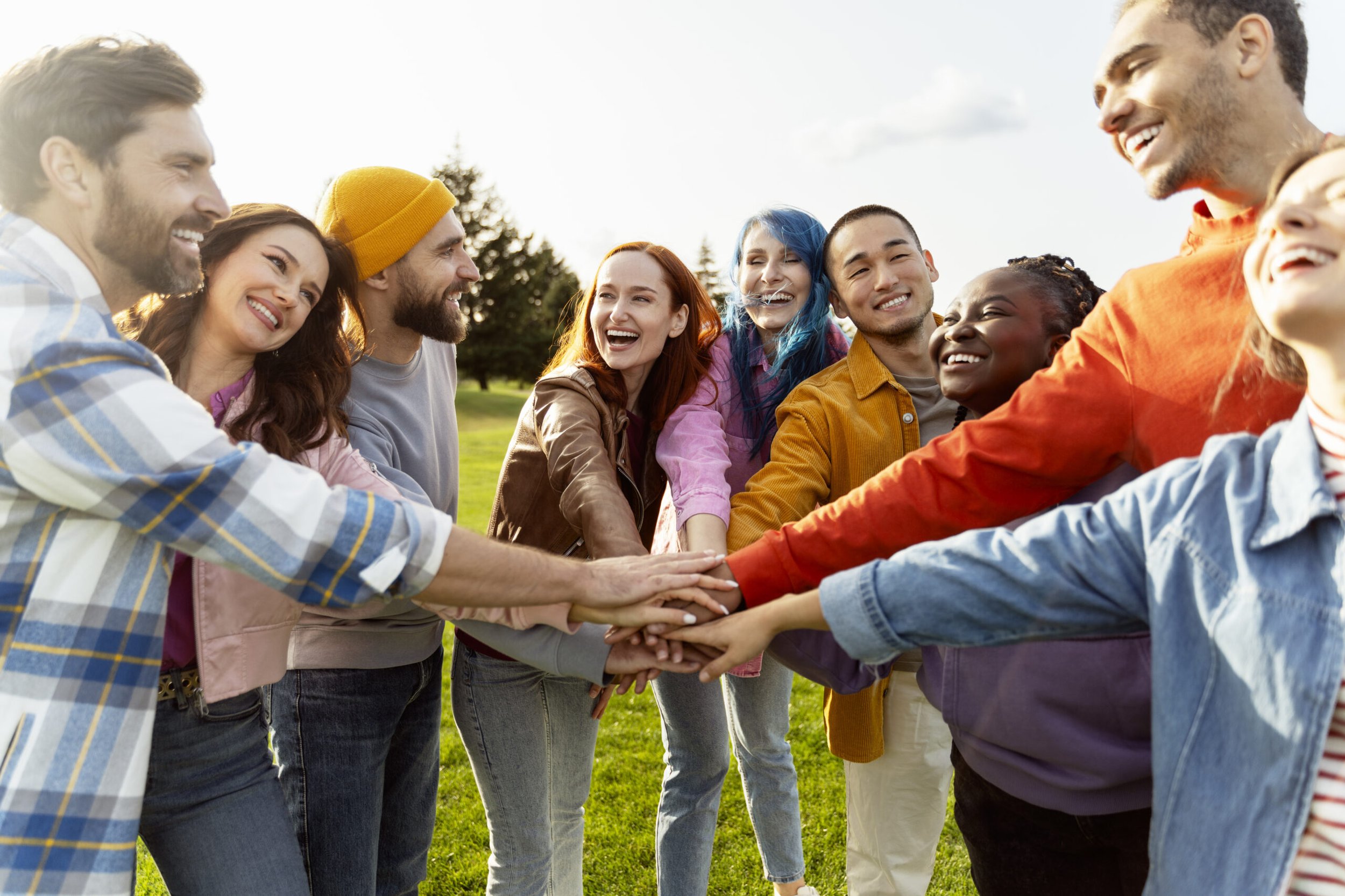 Group of diverse young adults outdoors, stacking hands together in a circle, smiling and laughing.
