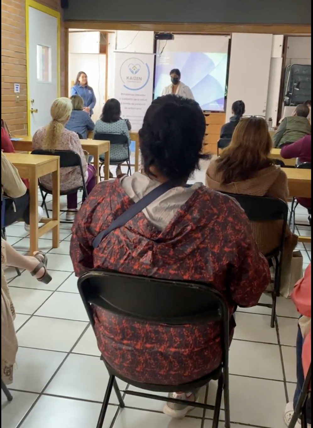 Personas asistiendo a una presentación en un salón, con una pantalla y un cartel que dice Kaizen.