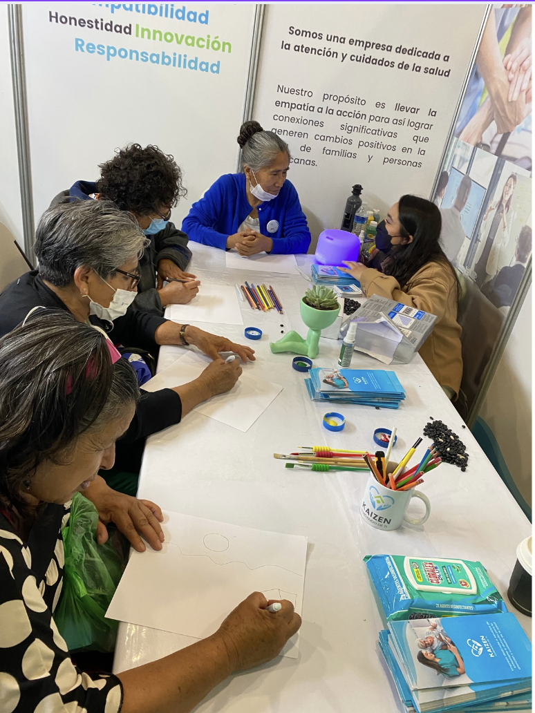 Varias personas sentadas alrededor de una mesa participando en un taller, algunos dibujando en hojas blancas, rodeadas de papeles, marcadores y flyers de una empresa dedicada a la atención y cuidado de la salud, con una pared en el fondo que tiene te