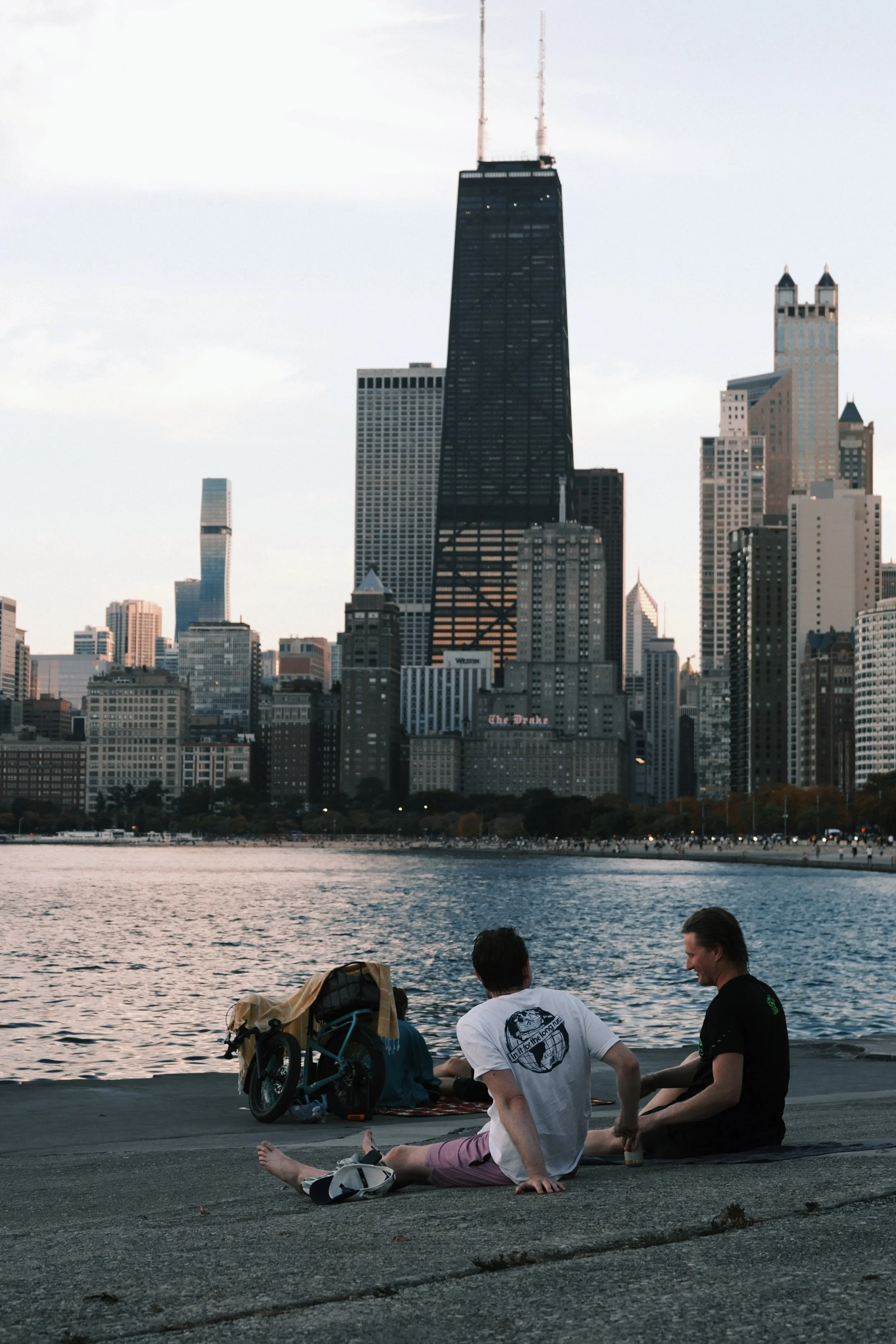 Two friends sitting on the lakefront steps beside a bike, skyline behind
