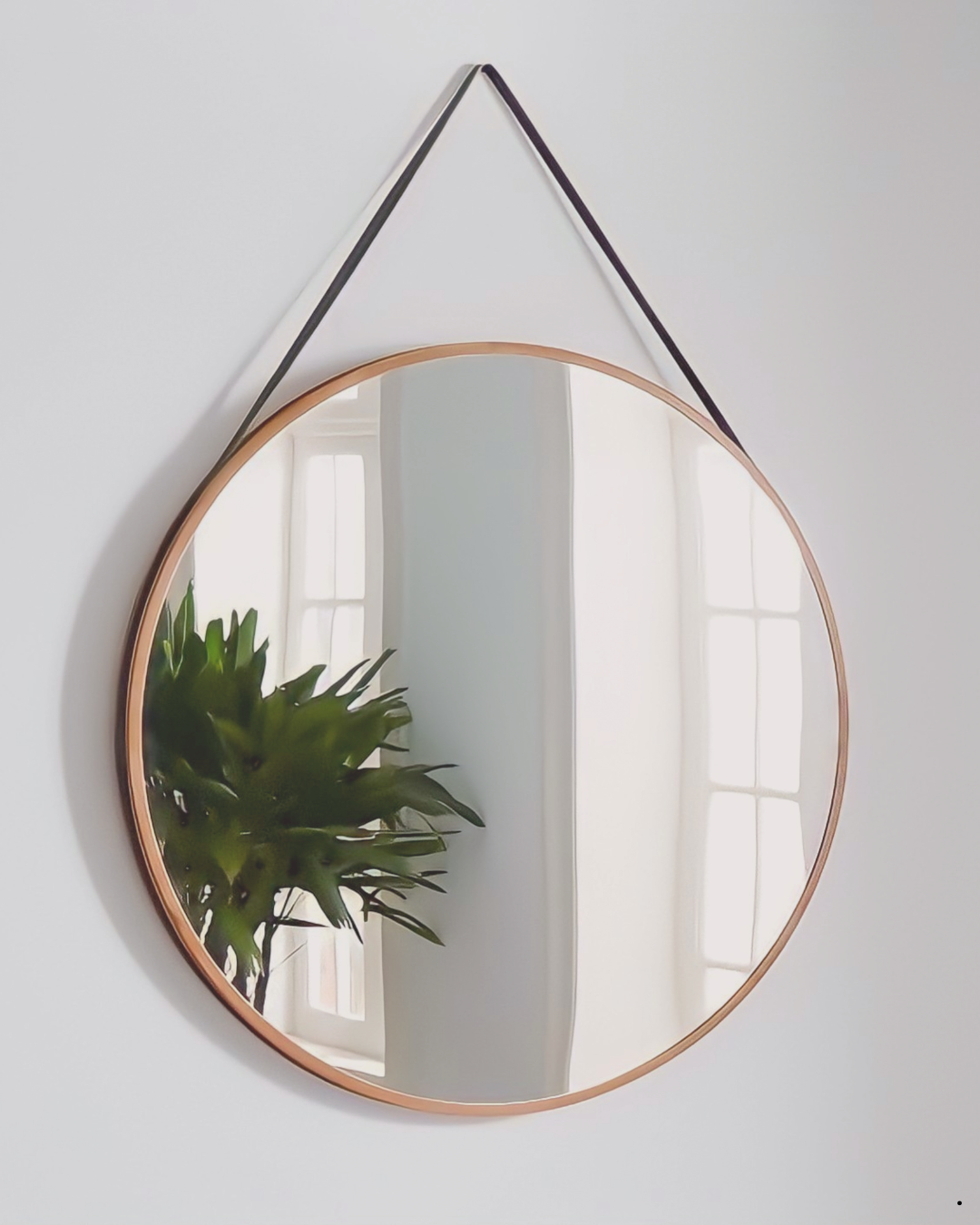 A round mirror hanging on a white wall with a leather strap, reflecting a window with light coming through and a green plant beside it.