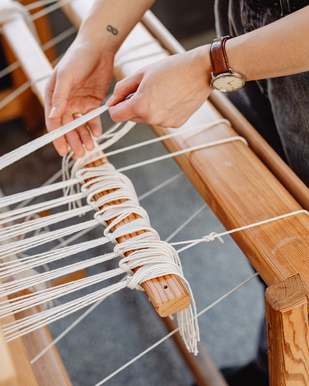 Person weaving white threads on a wooden loom, with hands adjusting the threads, wearing a watch and a ring.
