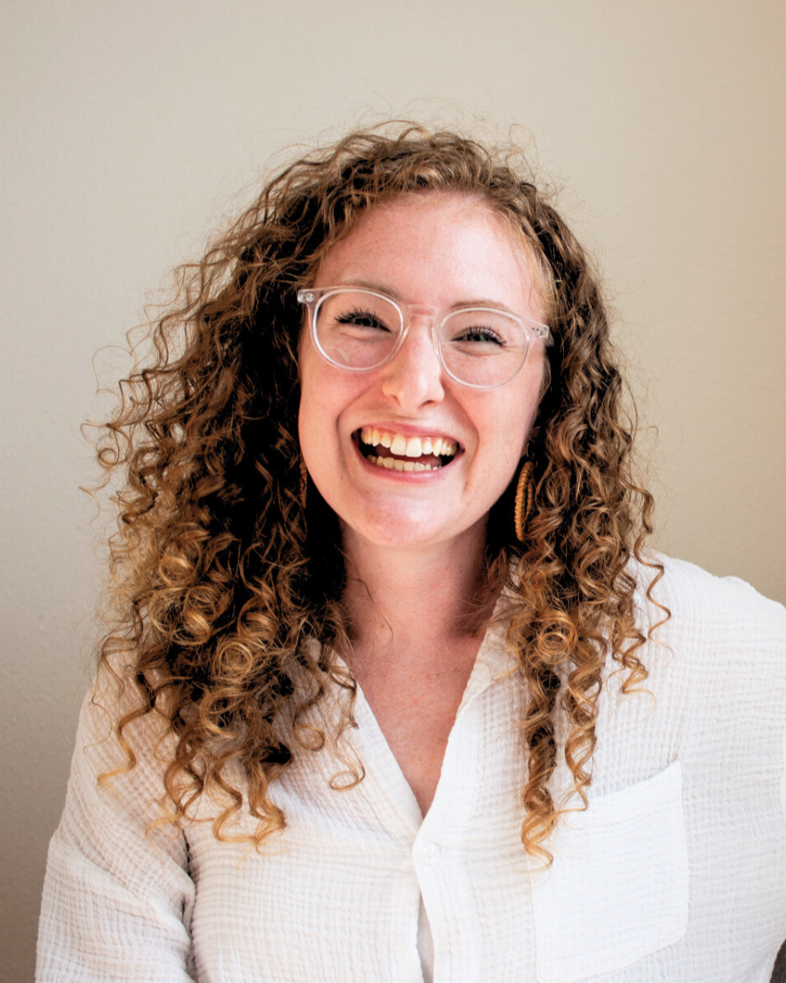 Woman with curly hair wearing glasses and a white shirt, smiling and showing teeth, against a plain background.