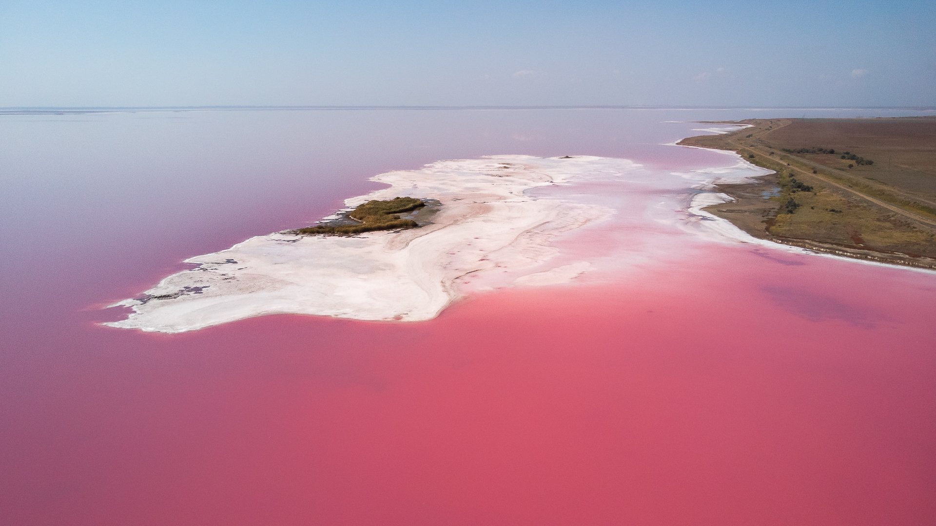 Aerial view of a pink salt lake with white salt deposits and a narrow strip of land on the right.