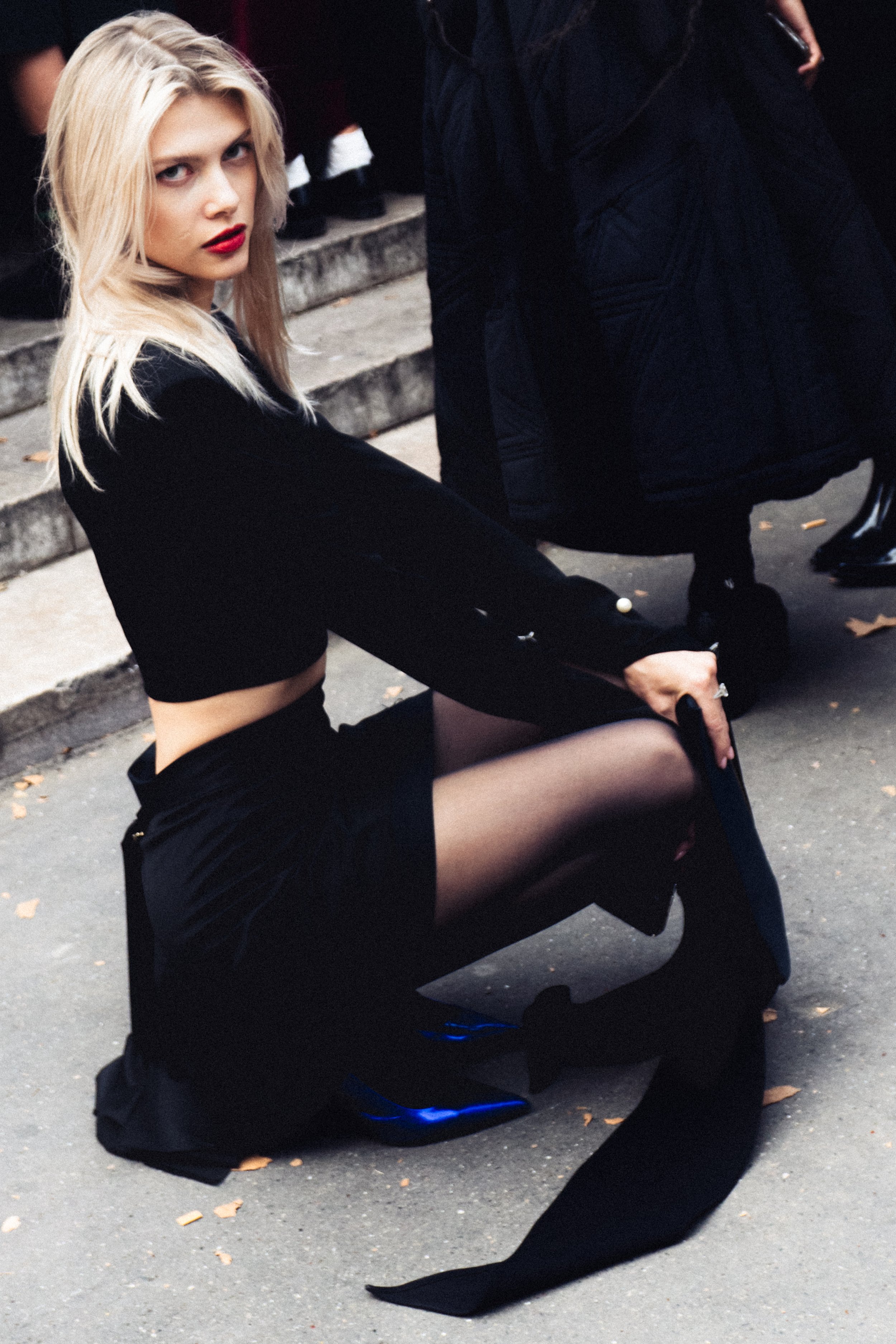 Fashion model squatting on the street, wearing a black outfit and blue shoes, looking at the camera. Vast Leaf