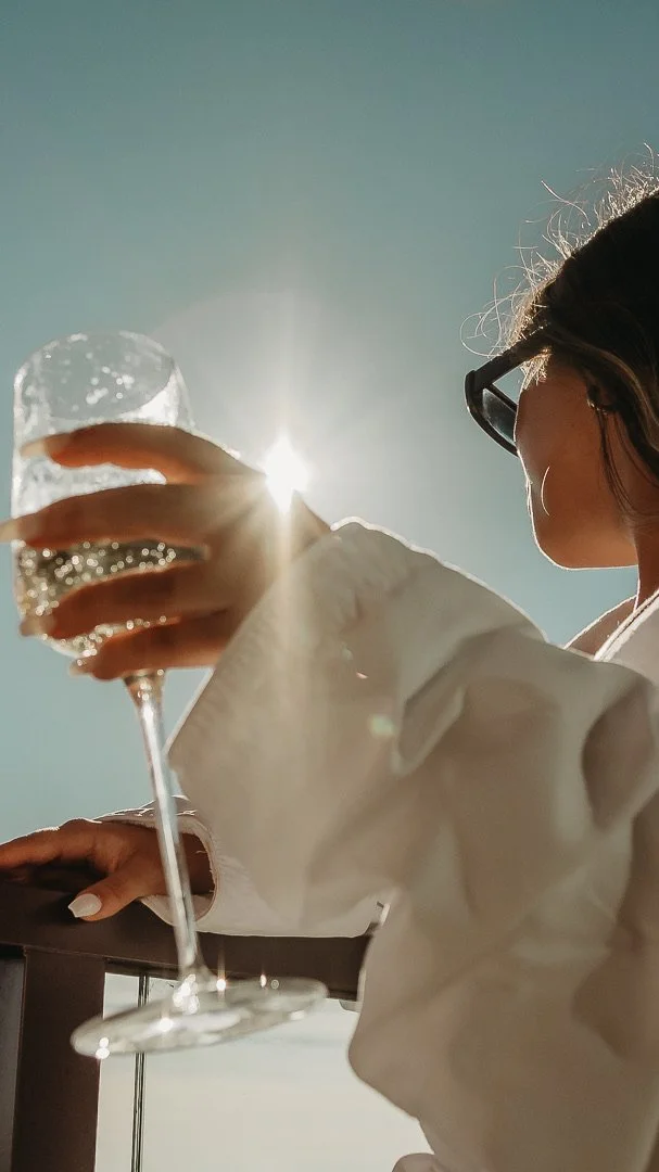 A woman with sunglasses enjoying a drink outdoors during daytime with the sun shining in the background.