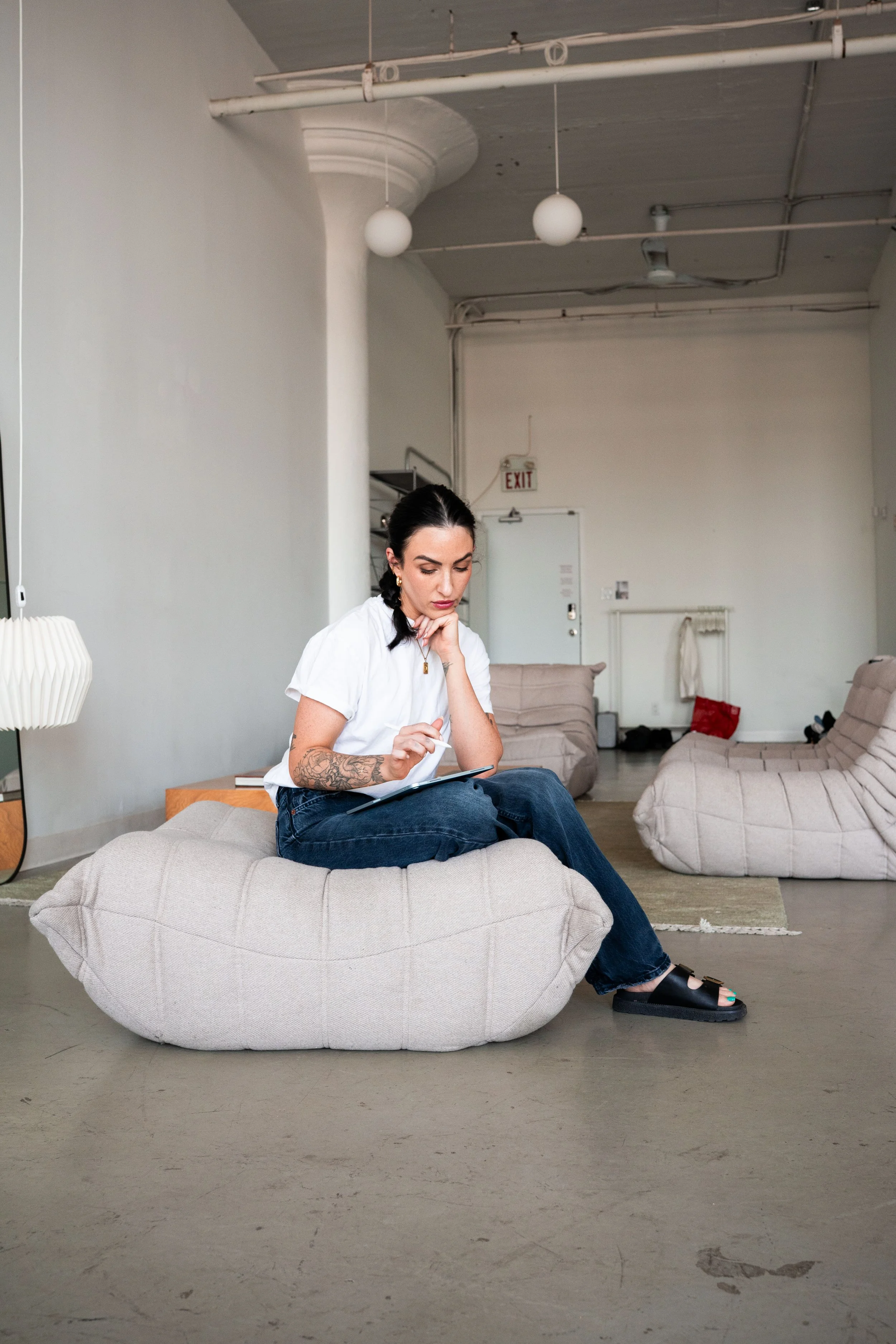 A woman with dark hair and tattoos on her arm is sitting on a beige bean bag chair in a spacious, industrial-style room. She is wearing a white t-shirt, dark jeans, and black sandals, and is writing on a notepad with a pen. The room has minimal furniture, including a side table with a lamp, and features some other bean bag chairs in the background. The walls are plain white, and the ceiling has exposed pipes and round light fixtures.