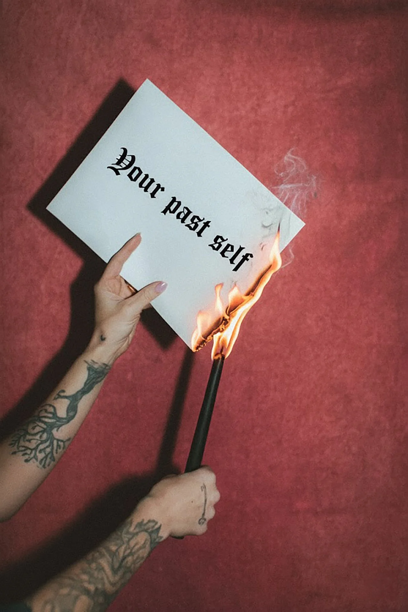 A hand with tattoos holds a lit match under a white piece of paper that reads 'Your past self' which is on fire against a red background.
