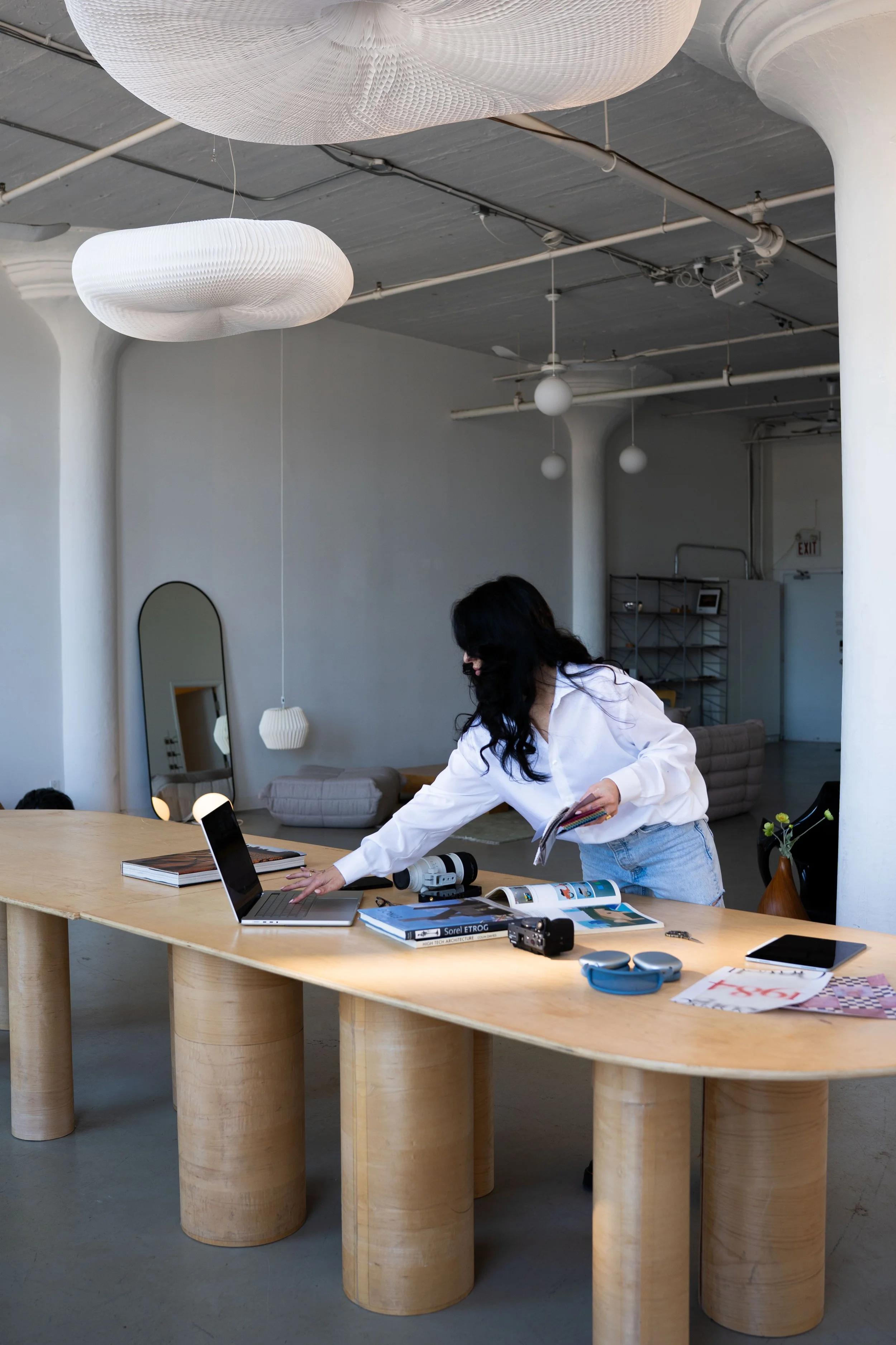 A woman with dark hair, dressed in a white shirt and light blue jeans, is reaching for a laptop on a large wooden table in a modern, minimalist interior. The table has various books, magazines, a camera, and electronic gadgets on it. The space features white walls, decorative hanging lamps, a mirror, and contemporary furniture.
