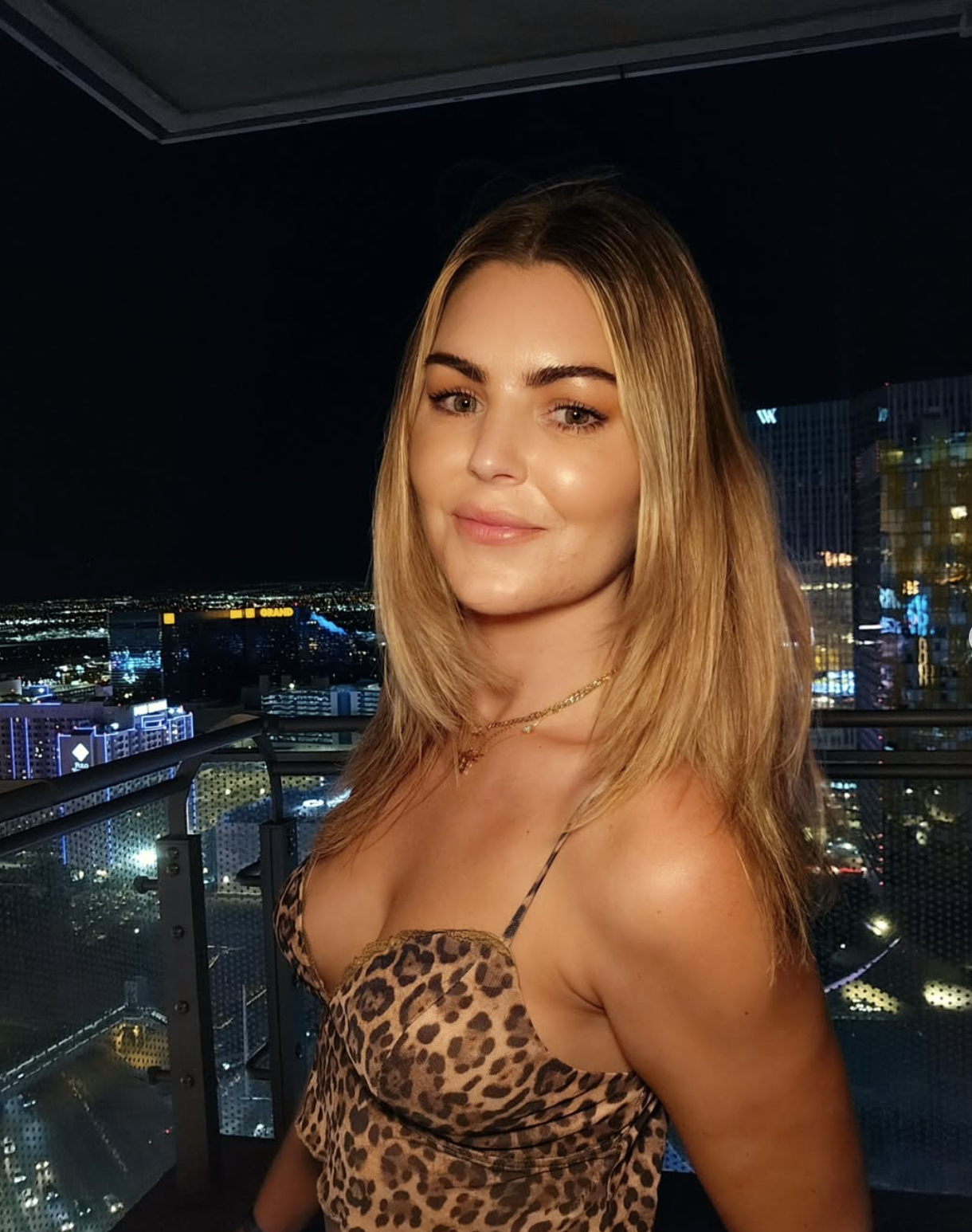 A young woman with blonde hair and light skin, wearing a leopard print top, posing on a balcony with a city skyline at night in the background.