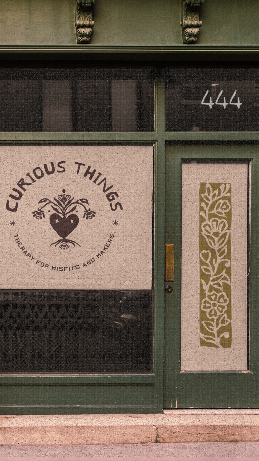 Green storefront window with a sign that reads 'Curious Things, Therapy for Misfits and Makers.' The sign has a heart with a plant growing out of it and an eye below. The window also has the number 444 written on it and a decorative floral pattern on