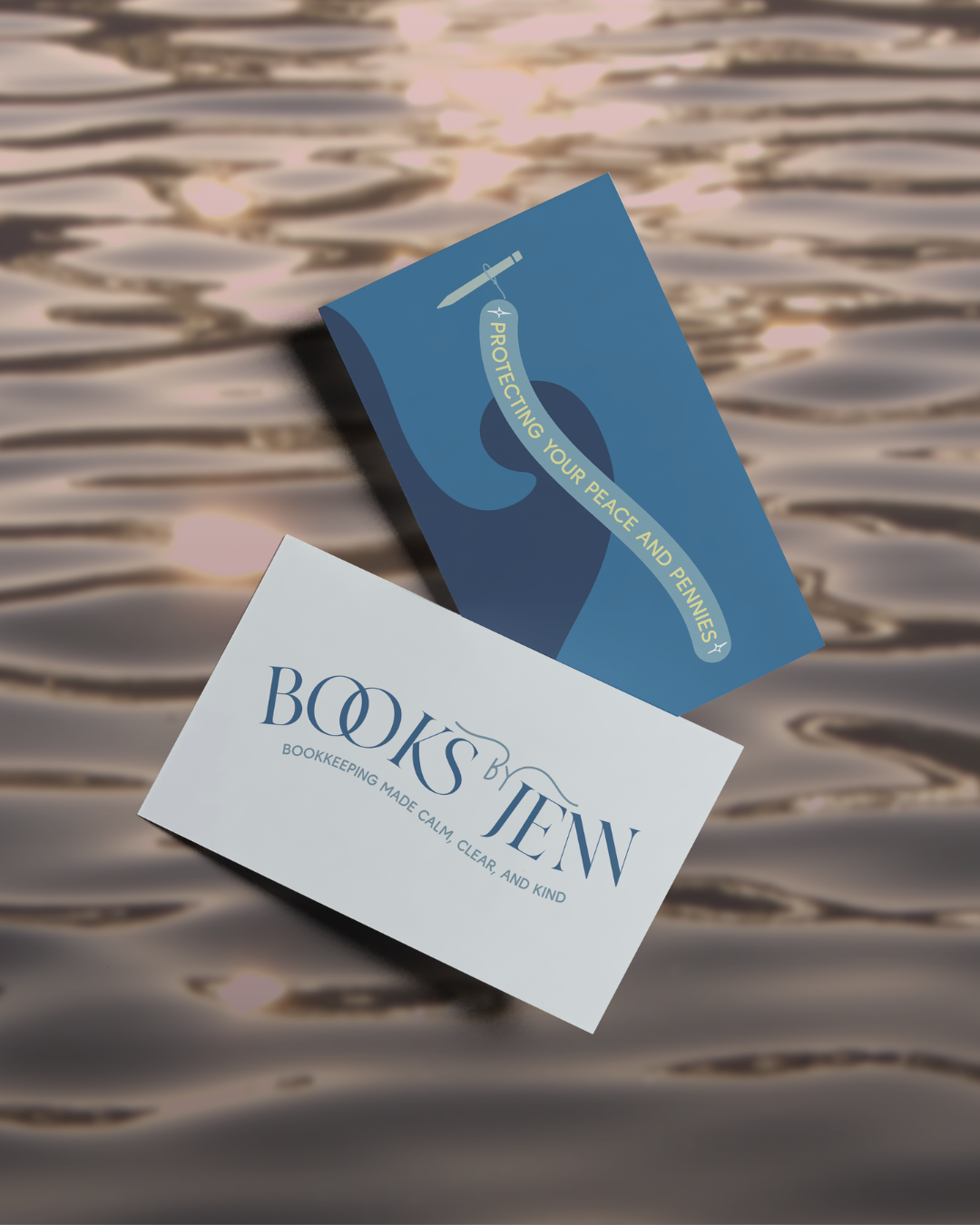 Two business cards floating on a water surface, one reading 'Books by Jen' with the tagline 'Bookkeeping made calm, clear, and kind,' and the other with a message about protecting peace and pennies, featuring a sword and ribbon design.