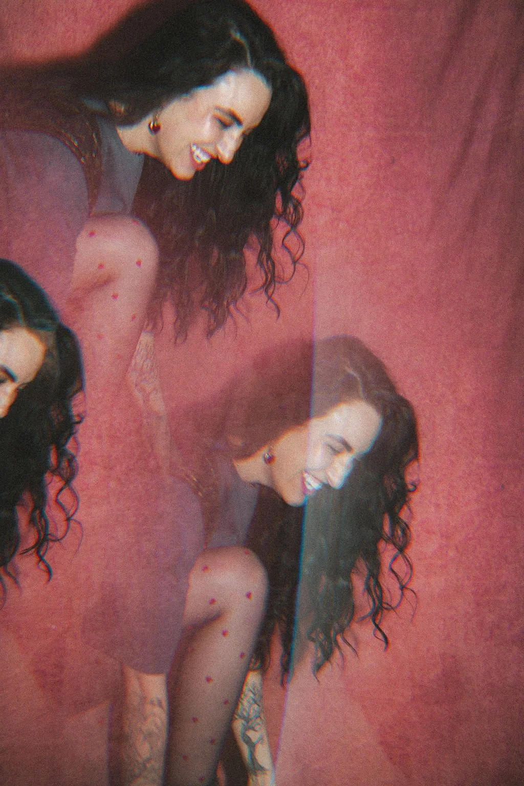 A woman with long curly hair, earrings, and a black dress, smiling and looking down against a pink wall.