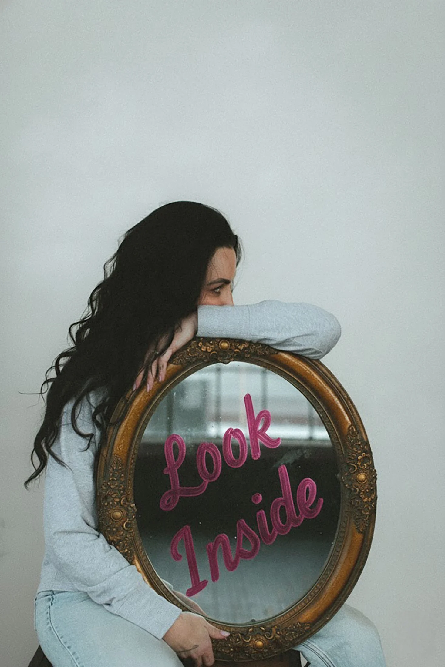 A woman with long black hair leaning on a vintage oval mirror with the words 'Look Inside' written in pink on the glass.