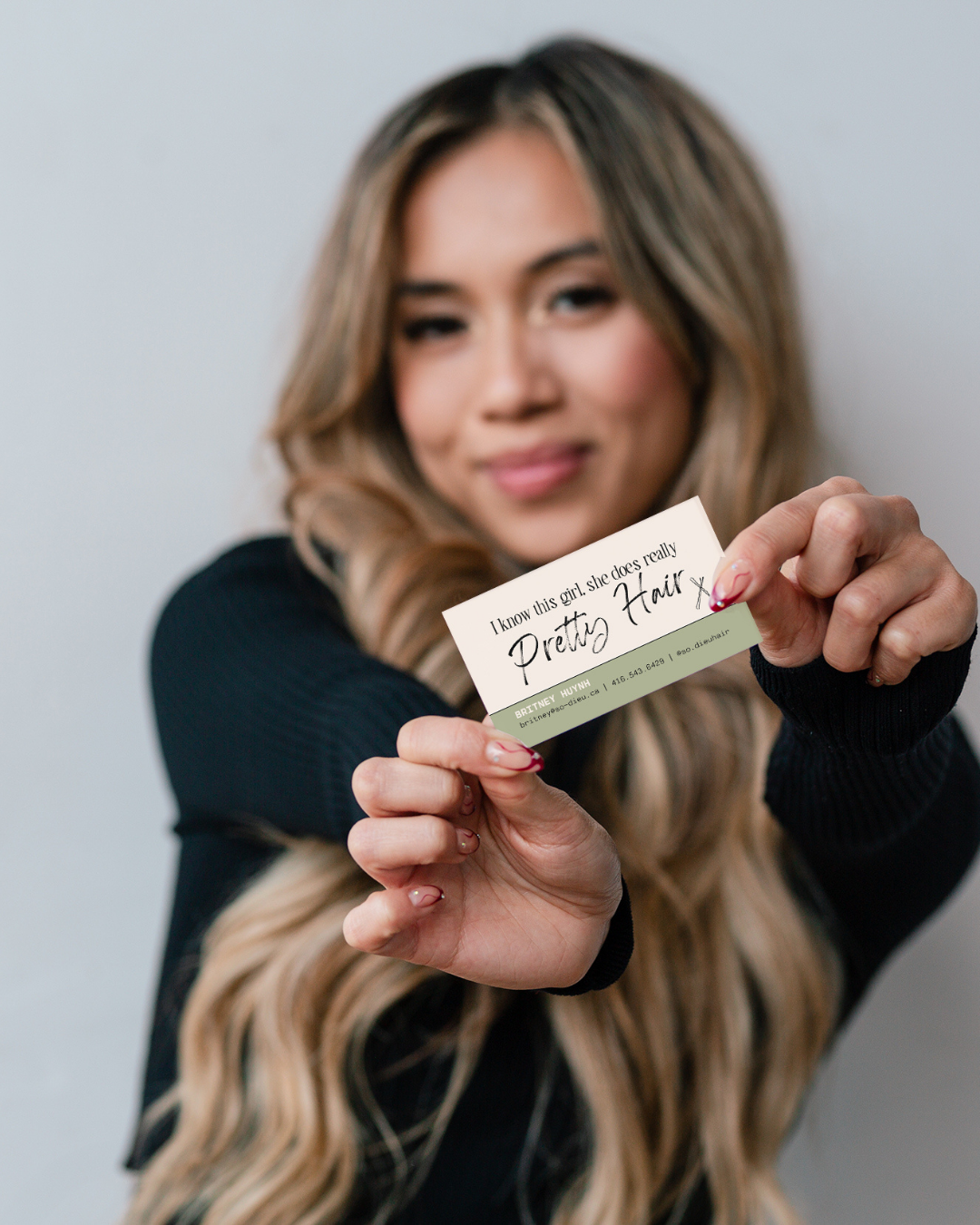 A woman with long, wavy blonde hair holding a business card for a hairstylist, smiling slightly, wearing a black top against a plain, light background.