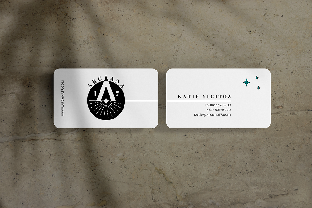 Two white business cards on a concrete surface, one with a black and white logo for Arcanai and the other with contact information for Katie YigitOz.