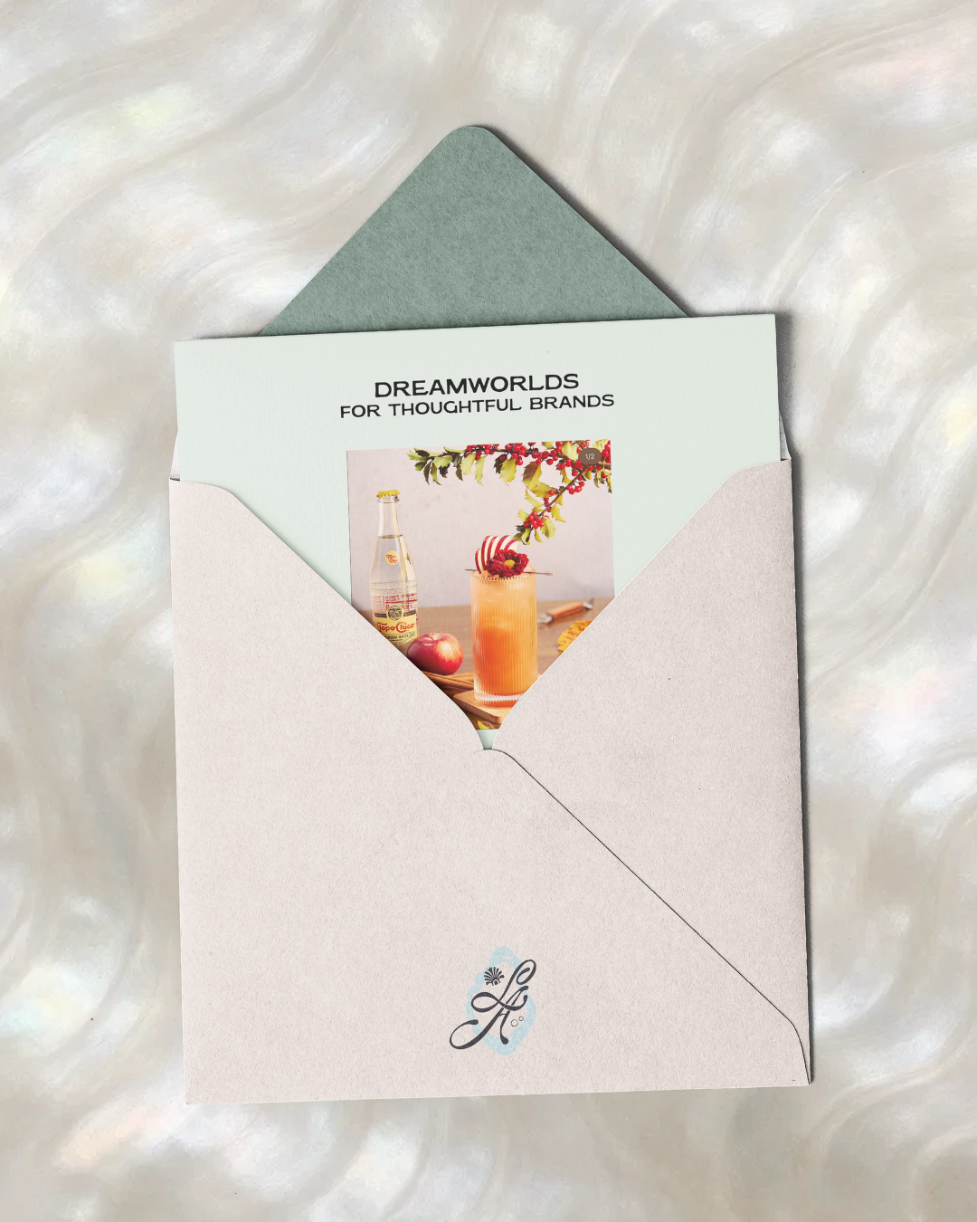 A white envelope with a blue interior flap and a decorative letter 'L' on the front, partially opened to reveal a card for 'Dreamworlds for Thoughtful Brands.' The card features a table with a drink garnished with berries and a branch, a bottle, and 