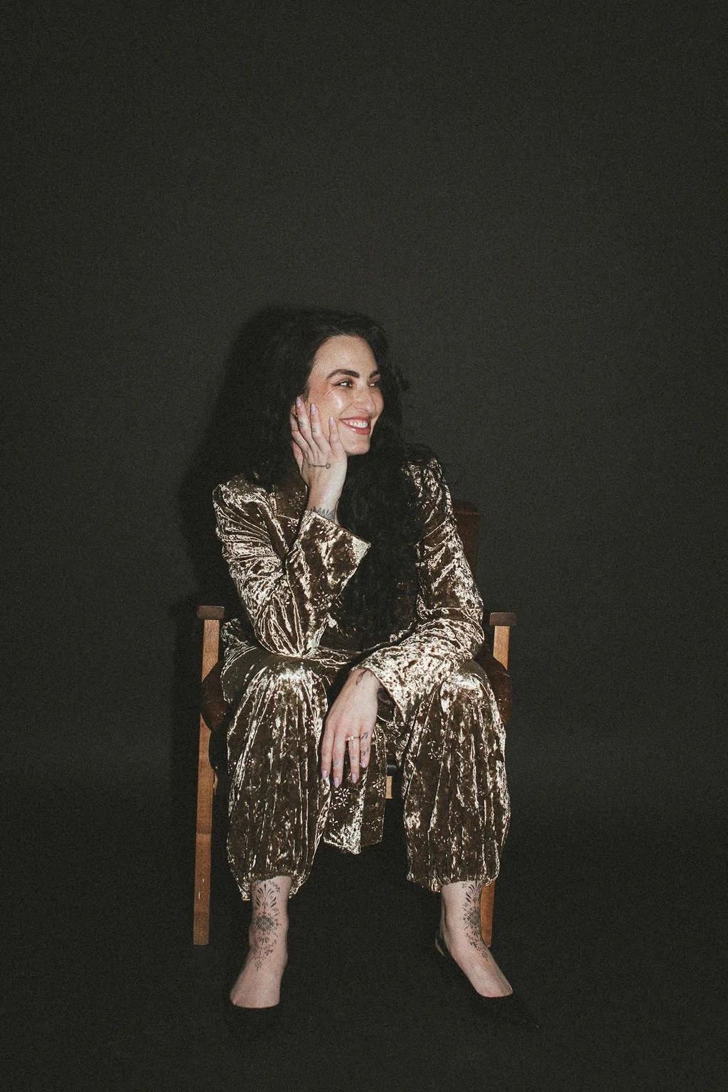 A woman with long dark hair sitting on a wooden chair, wearing a gold velvety suit, smiling and looking to the side against a dark background.
