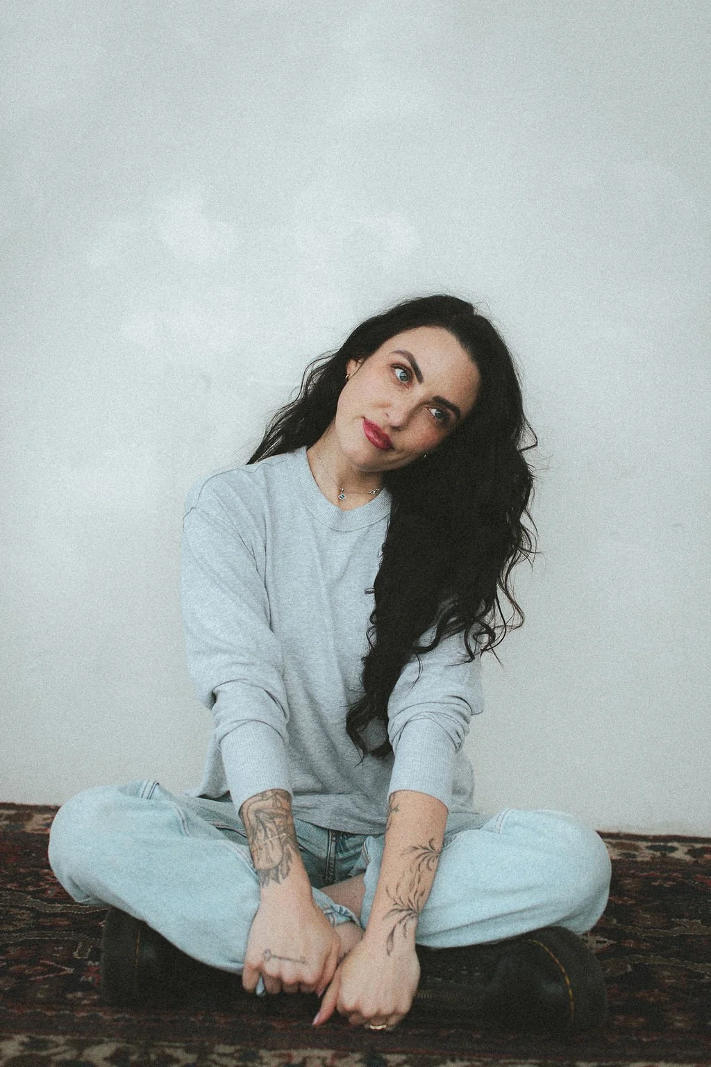 Brand photos of A young woman with long curly black hair, sitting cross-legged on a patterned carpet against a plain white wall, wearing a light gray sweatshirt and ripped jeans, with tattoos on her arms, looking at the camera with a slight smile.