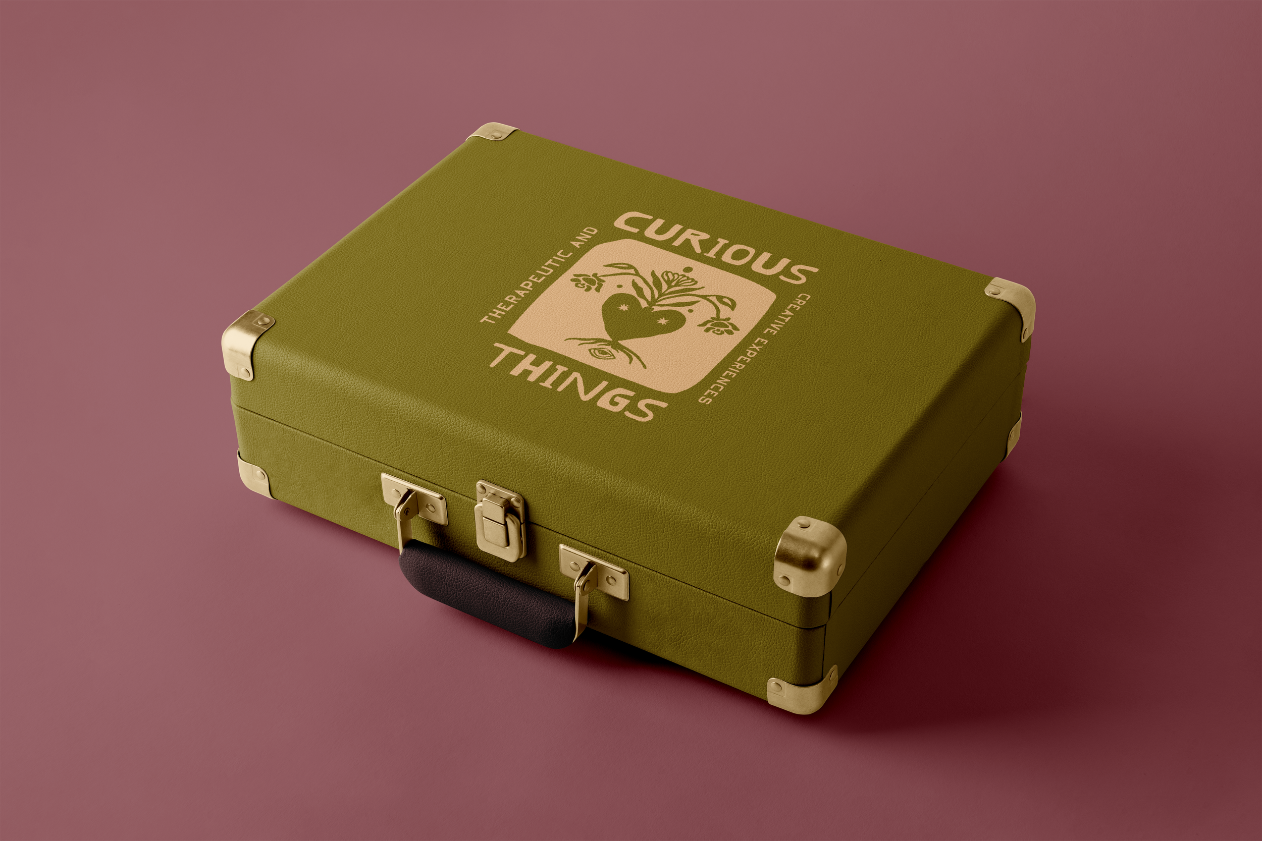 A green vintage-style briefcase with metal corners and latch, featuring a graphic of a heart with leaves and the text 'Curious Things: The Artistic and Real-Life Experiences' on the cover, set against a mauve background.