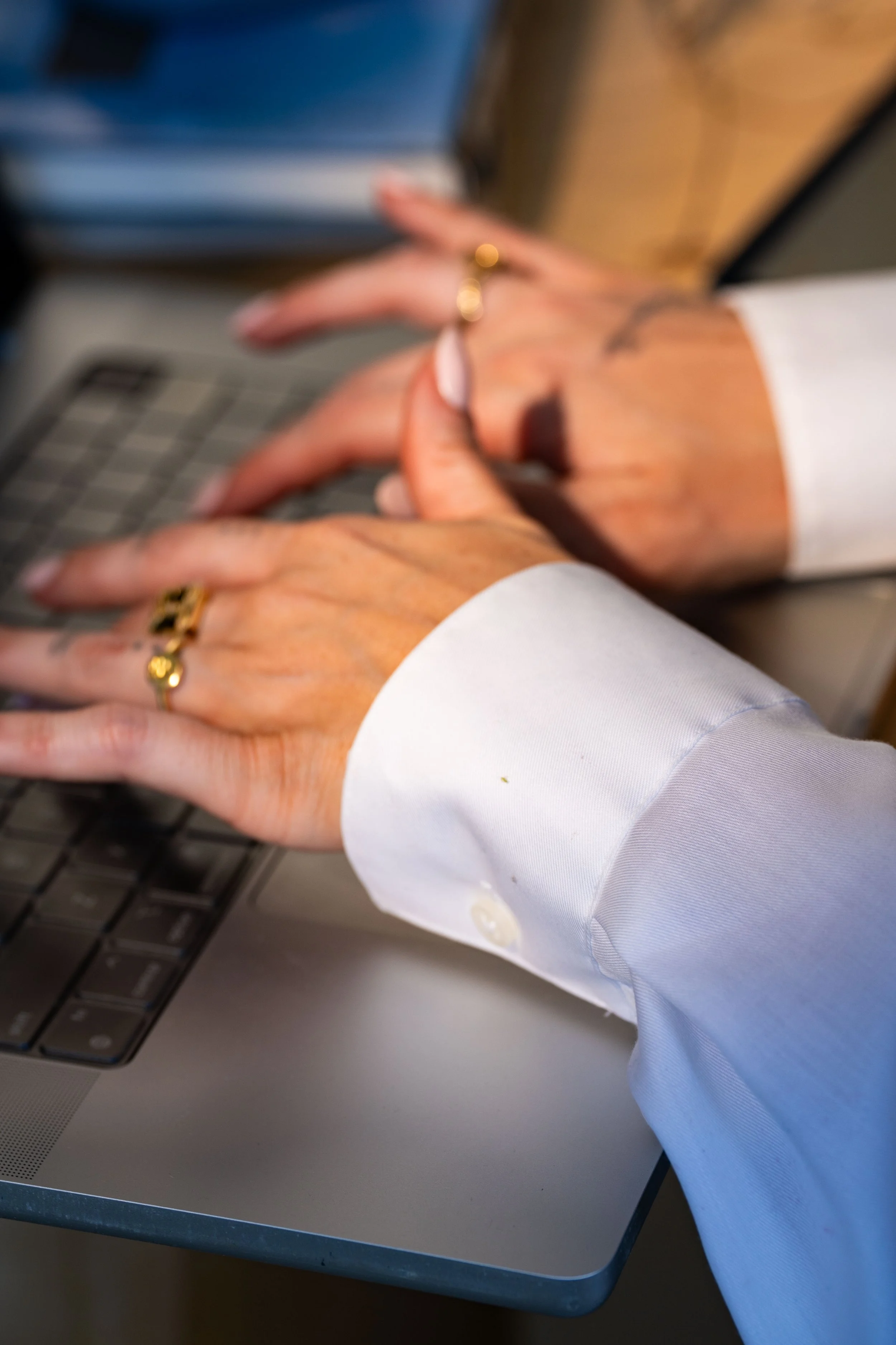 Close-up of two hands with rings resting on a laptop keyboard, wearing white shirts.