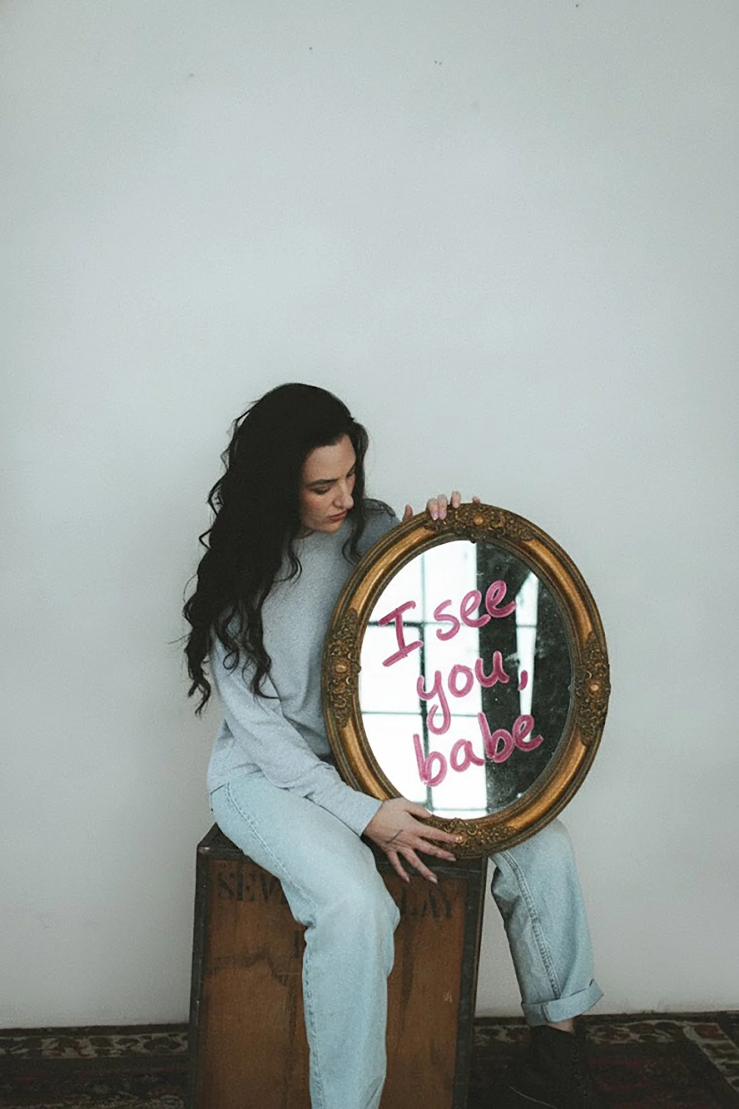 A woman with long dark hair sitting on a wooden box holding an ornate oval mirror with the words 'I see you, babe' written on it in pink