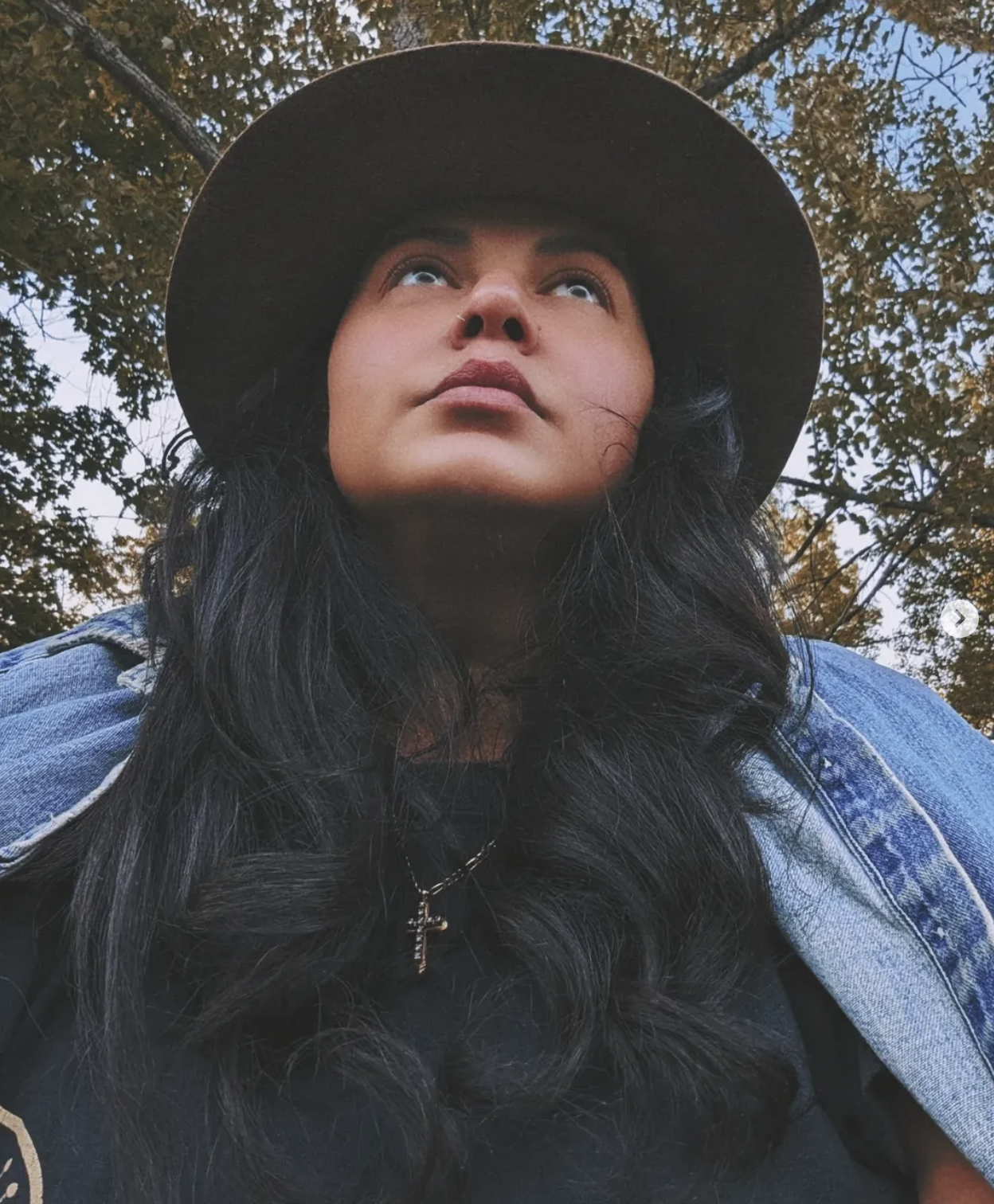 A woman with long black hair wearing a black hat and a black shirt, looking upward with trees and sky in the background.