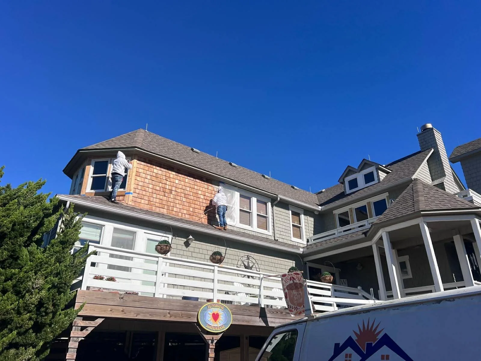 Two workers on the roof of a large, multi-story house painting or cleaning the exterior walls, with a clear blue sky in the background.