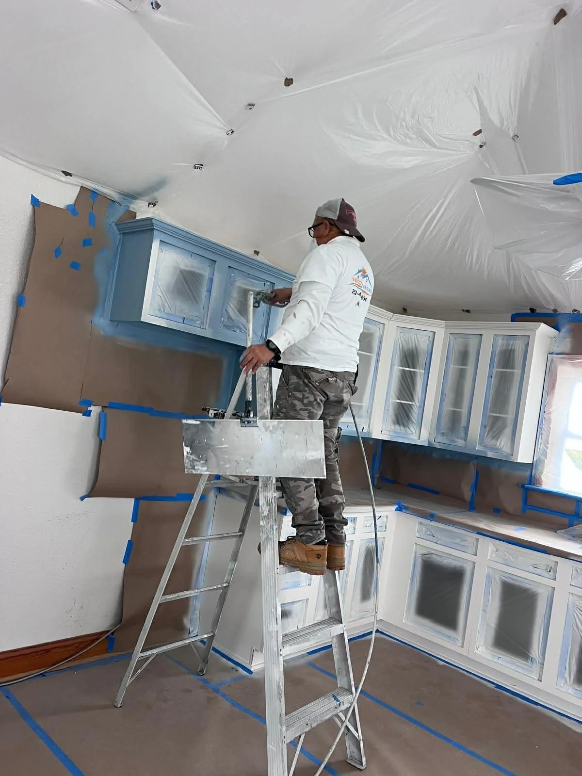 A person is standing on a ladder, spray painting kitchen cabinets in a room covered with protective sheets and tape.