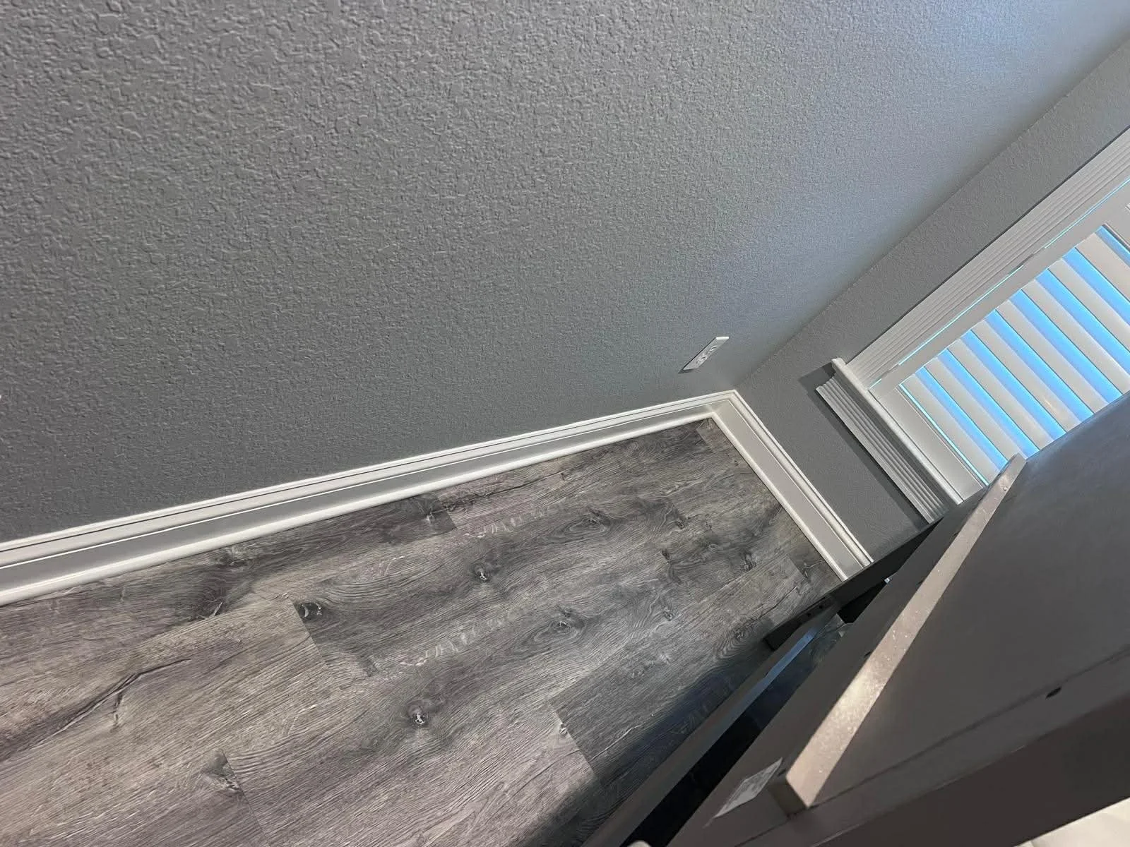 Interior corner showing textured gray wall, white baseboard, dark gray wood-look flooring, gray window blinds, and part of a black chair.