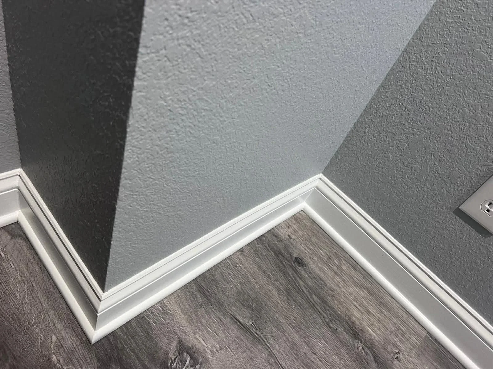 Close-up of a corner in a room showing baseboards, a textured gray wall, and part of an electrical outlet.