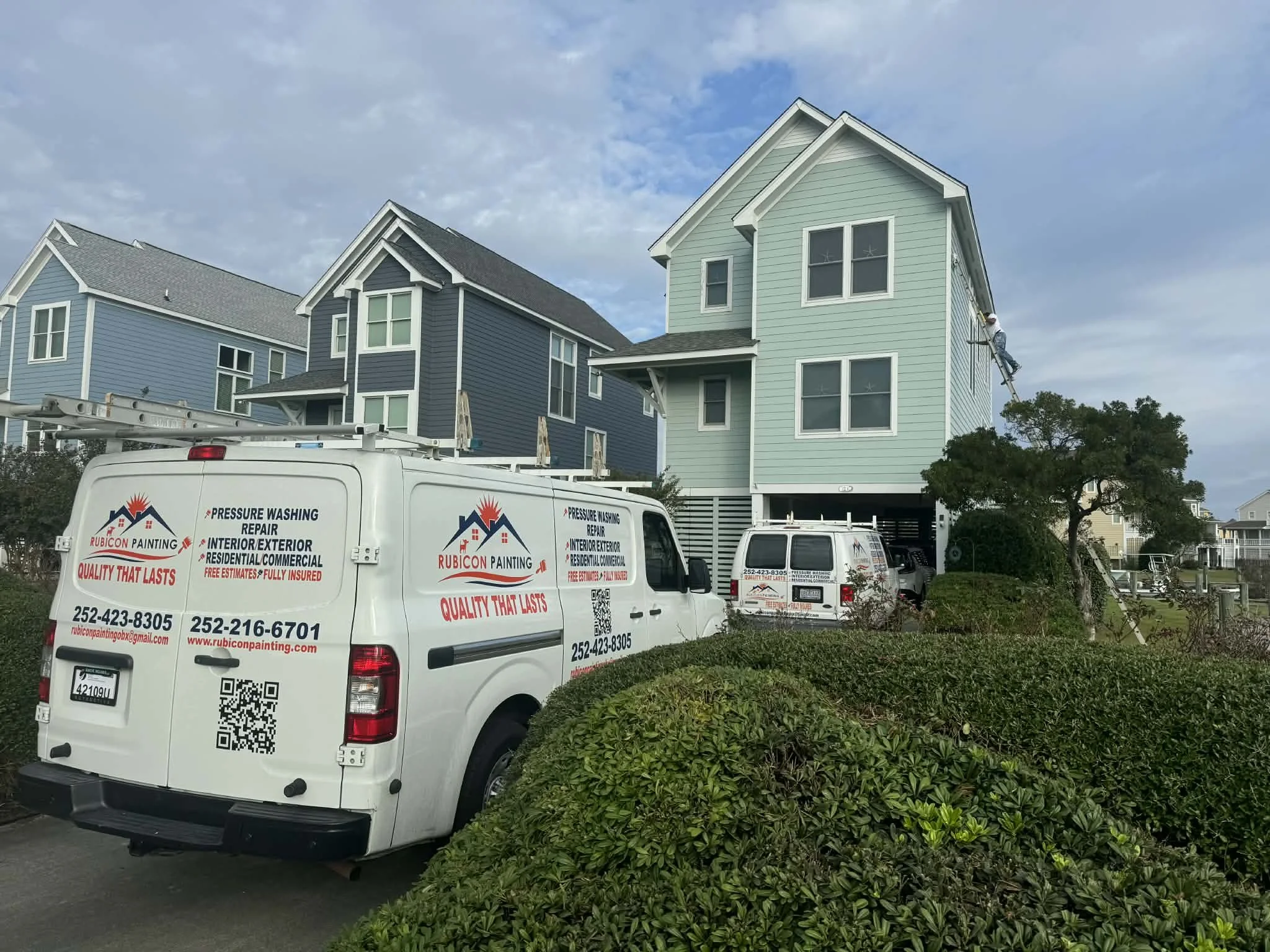A house painter on scaffolding painting exterior of a light green, three-story house. A white company van with painting and repair services parked nearby, branded with 'Rubicon Painting' and contact information.