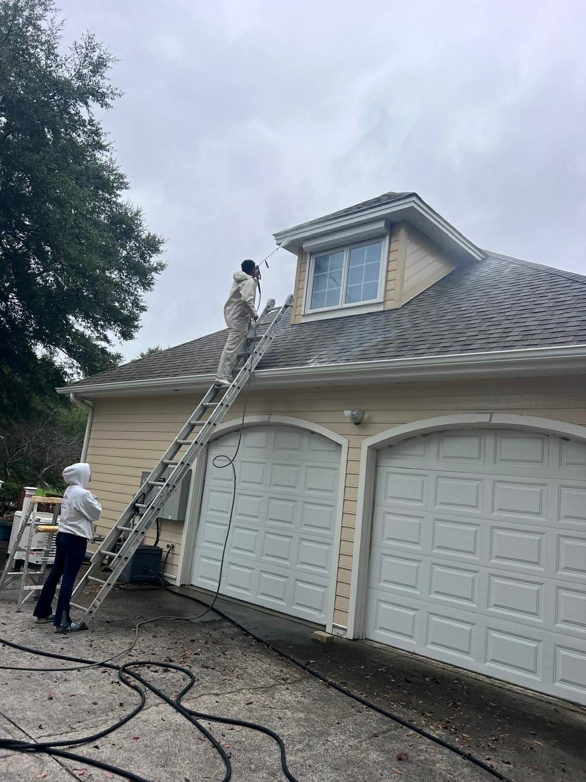 Person cleaning or inspecting the roof of a house while standing on a ladder, with another person observing nearby on a cloudy day.