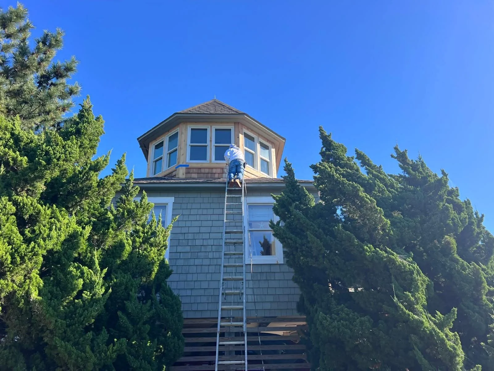 A person on a ladder working on the top of a house with a turret, surrounded by trees and set against a bright blue sky.