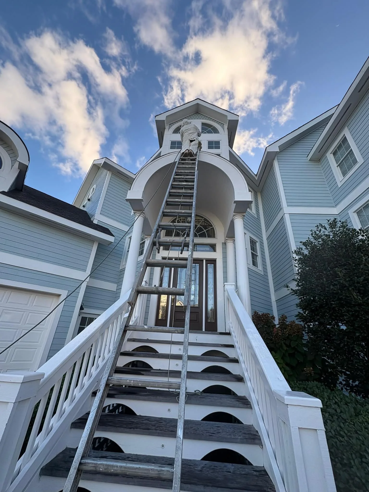 A person on a ladder working on the exterior of a large, multi-story house with light blue siding, white trim, and a dark front door, against a backdrop of a partly cloudy sky.