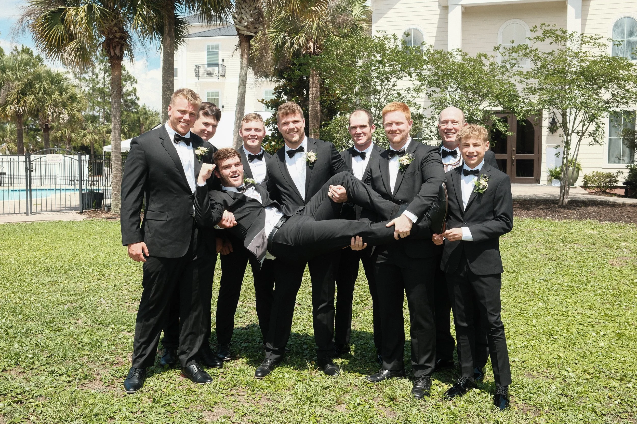 Group of nine young men dressed in black tuxedos with bow ties, some wearing white shirts and boutonnières, are outdoors on grass with palm trees and a white building in the background. One man is being held horizontally by the others, smiling and ma