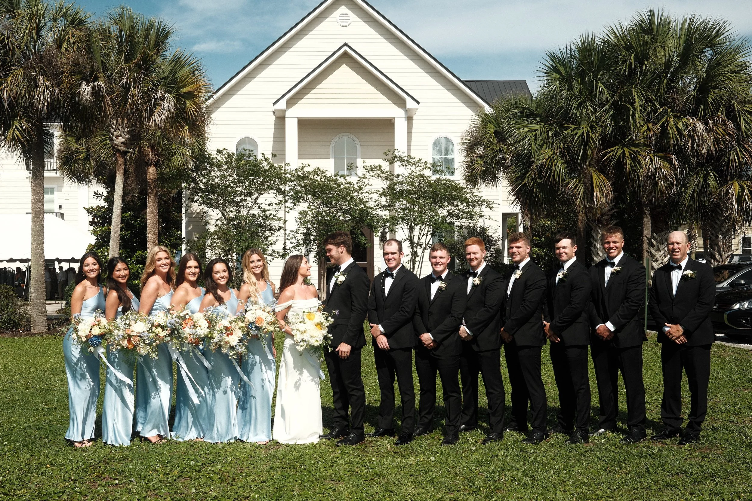 A group of people dressed in formal attire, standing outdoors on grass in front of a white house with palm trees, during a wedding celebration.