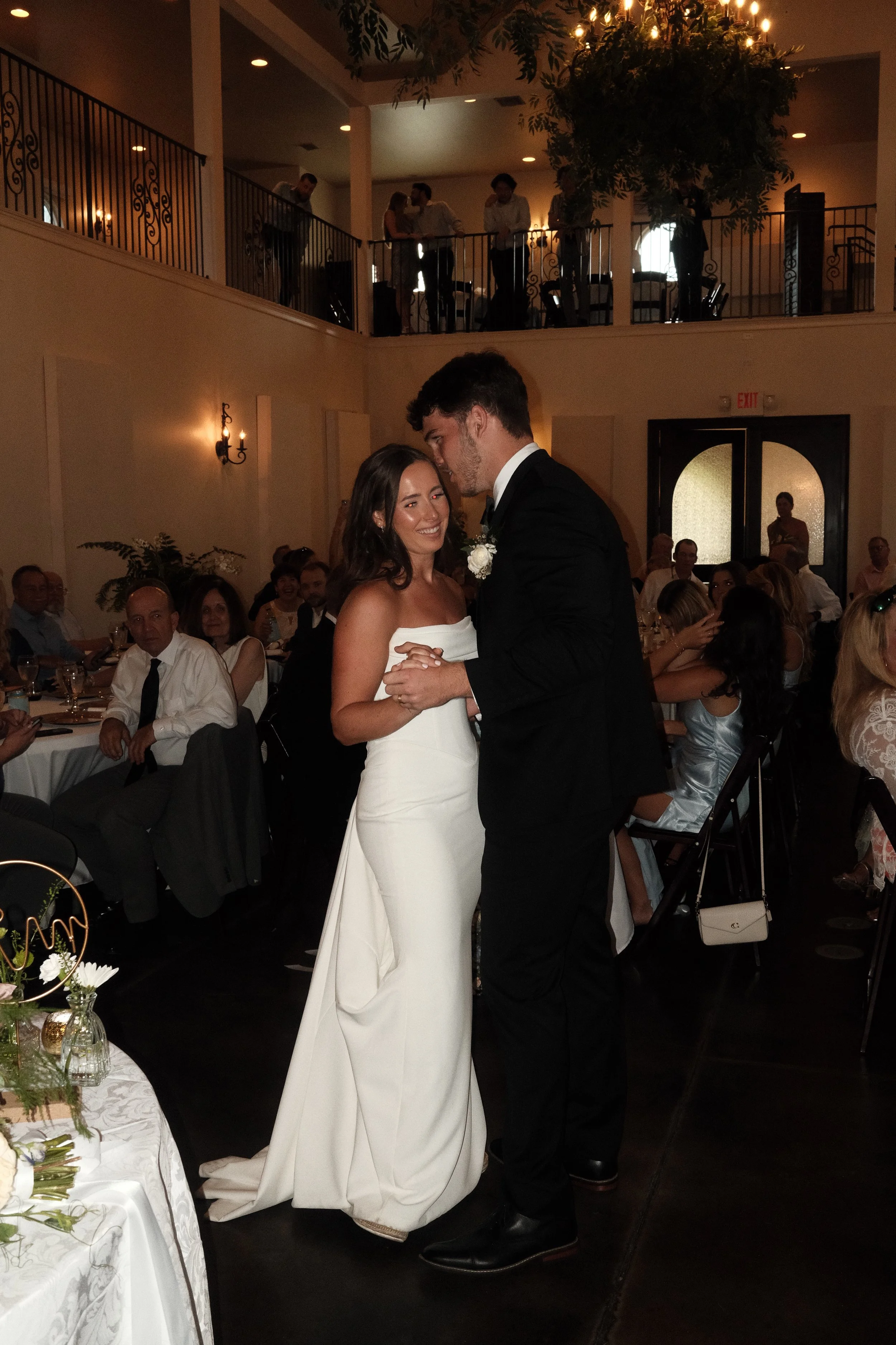 A bride and groom are sharing a dance at their wedding reception. The bride is wearing a strapless white wedding gown, and the groom is dressed in a black tuxedo. The wedding guests are seated at tables in the background, and some people are standing