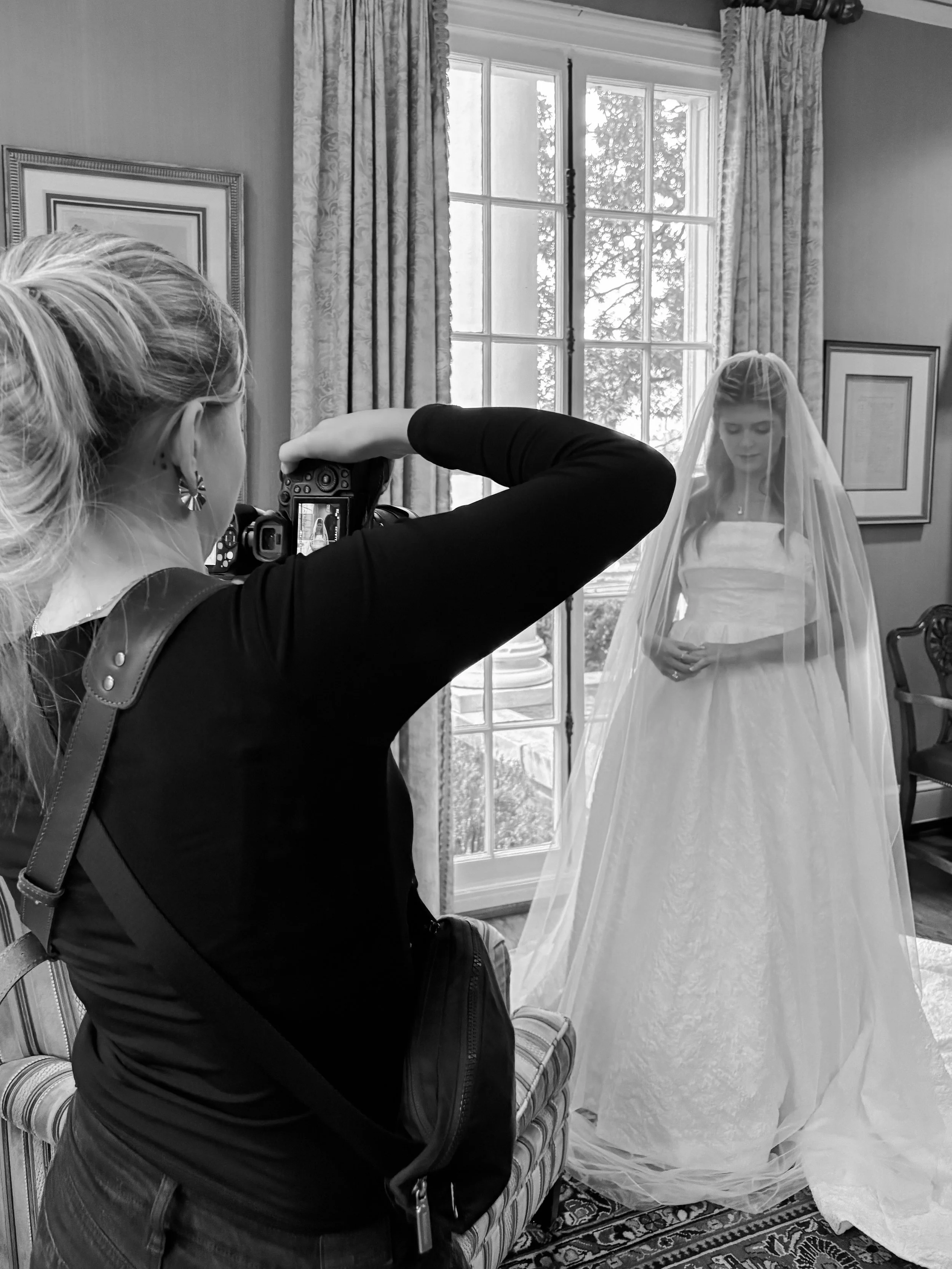 A photographer taking a photo of a woman dressed in a wedding gown and veil indoors next to a window.