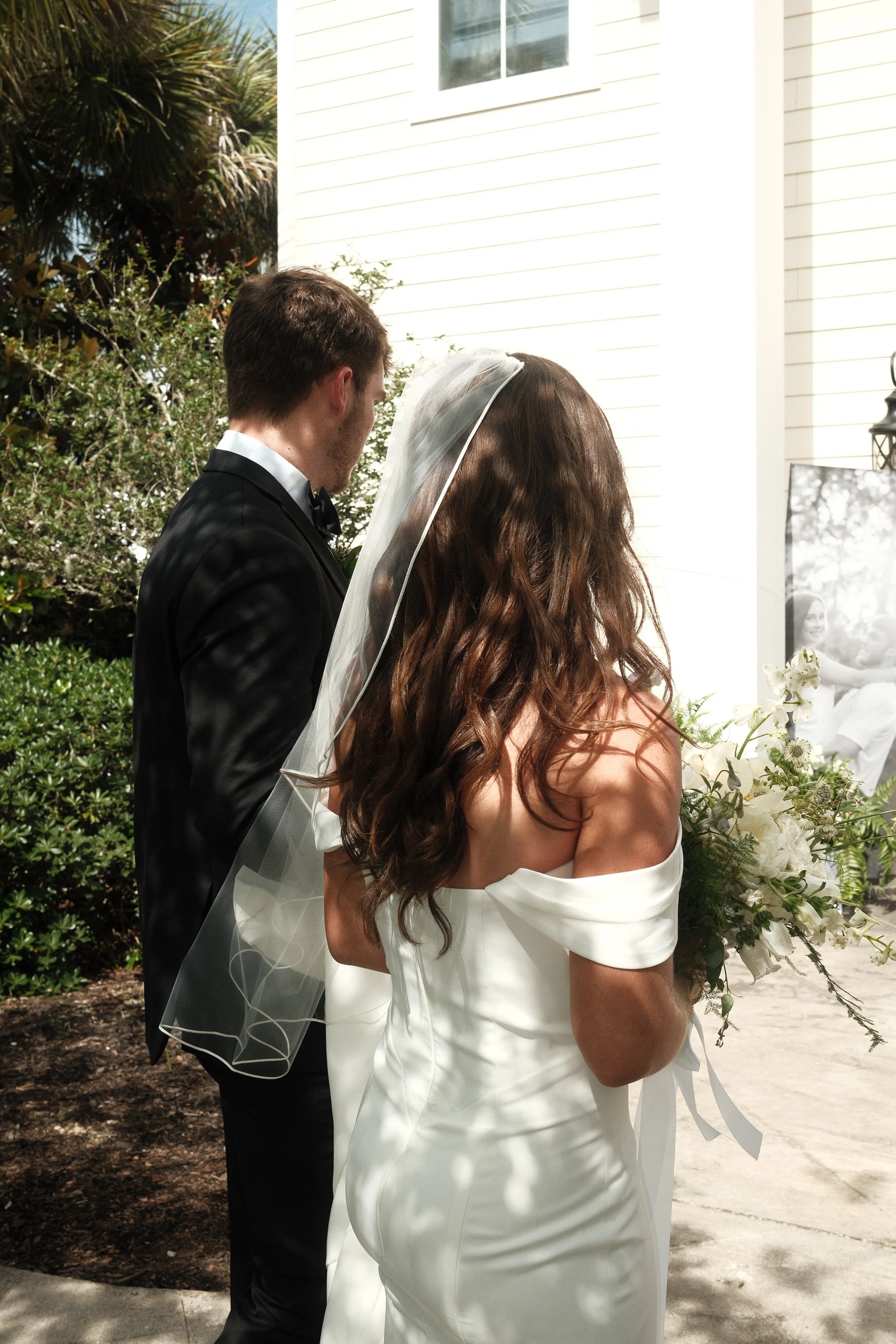 A bride and groom standing outdoors during a wedding ceremony, facing each other with their backs to the camera. The groom is wearing a black tuxedo and the bride is dressed in a white gown with a veil and long, wavy brown hair. There are flowers and